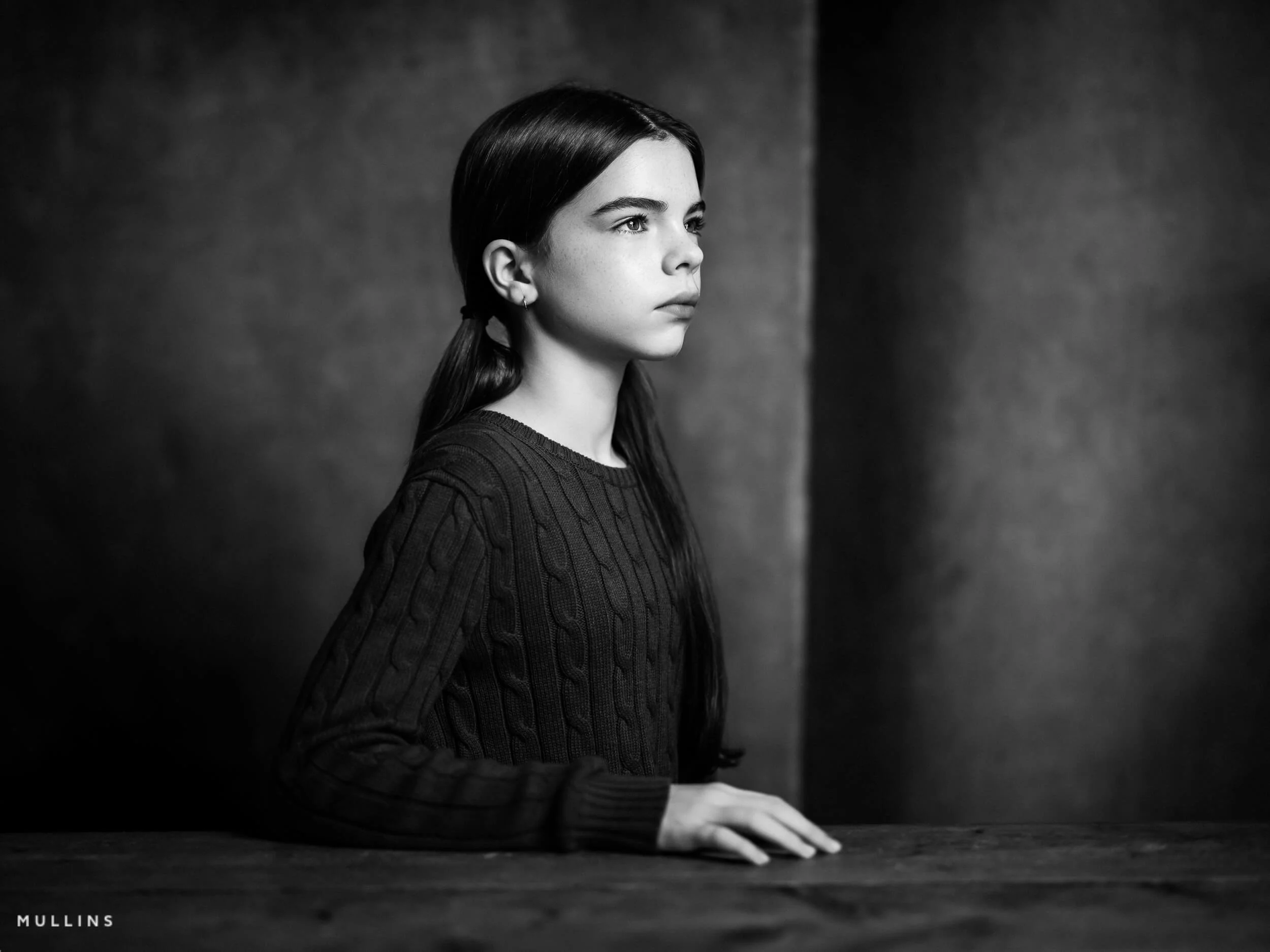 Black and white side-profile portrait of a young female actor seated at a wooden table in the studio.