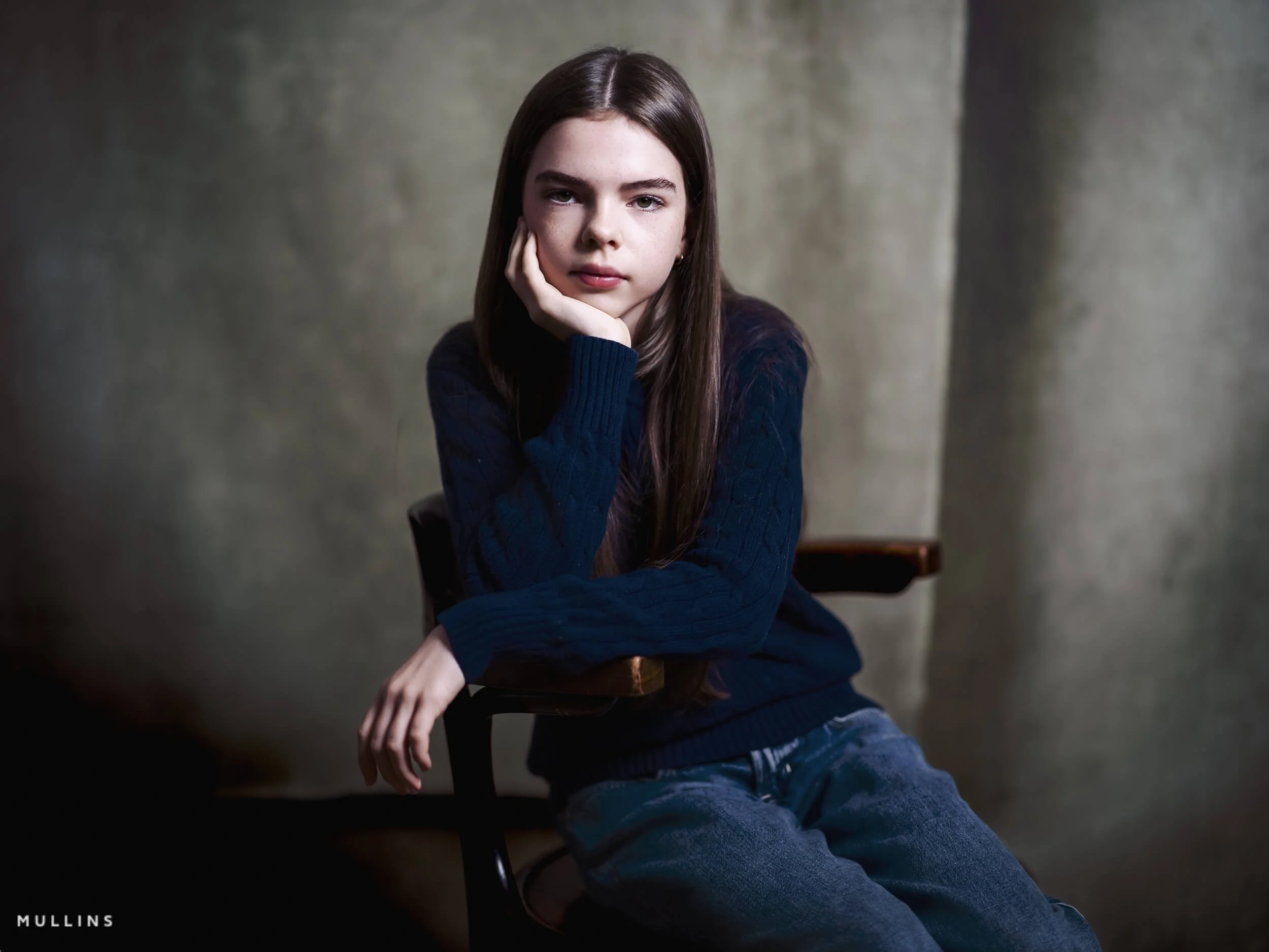 Seated portrait of a young female actor leaning on one hand in the studio, wearing a dark jumper and jeans.