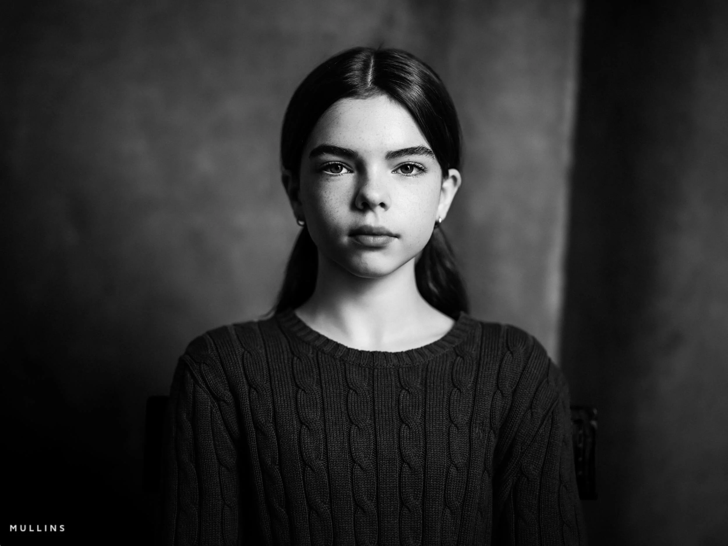 Black and white close portrait of a young female actor in a jumper, seated against a soft studio background.