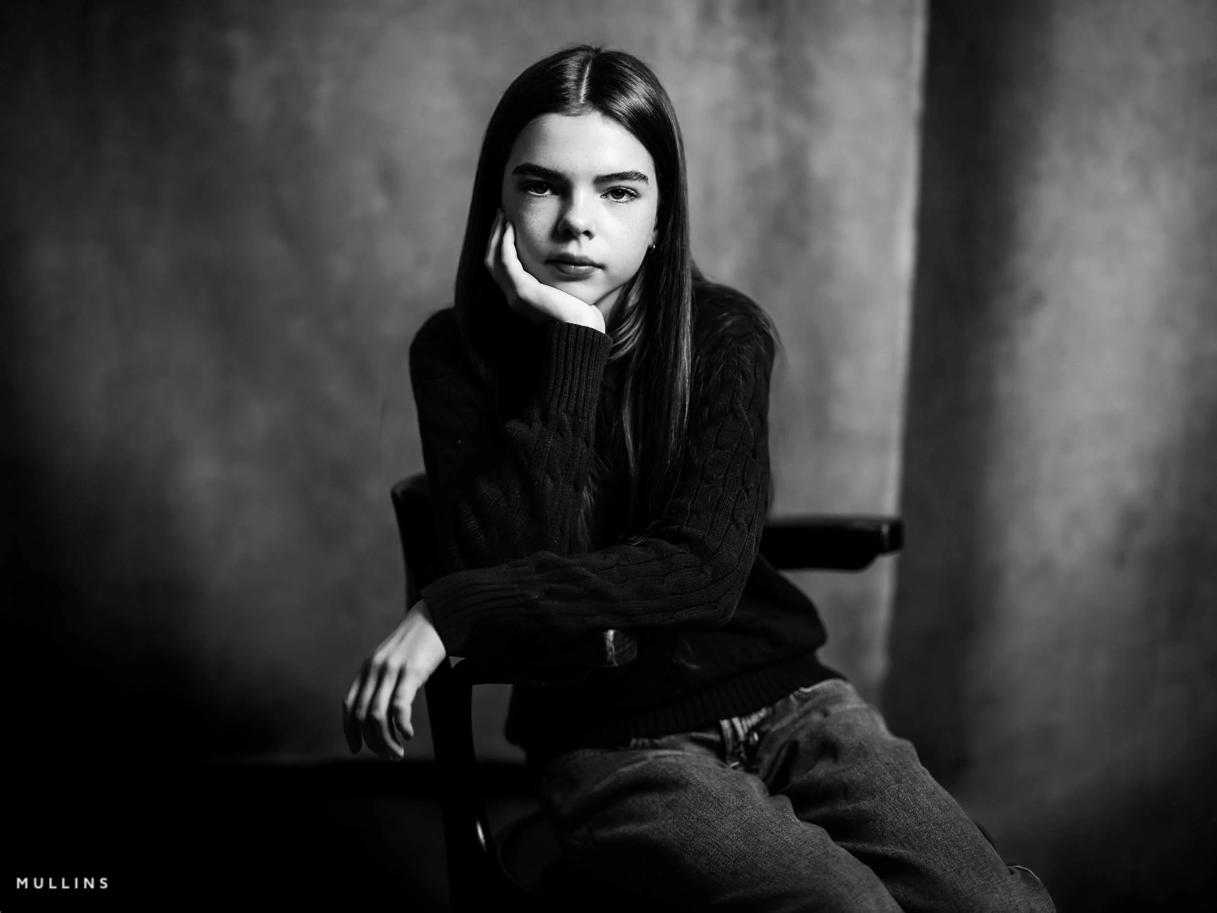 Black and white seated portrait of a young female actor leaning on one hand, wearing a dark jumper and jeans.
