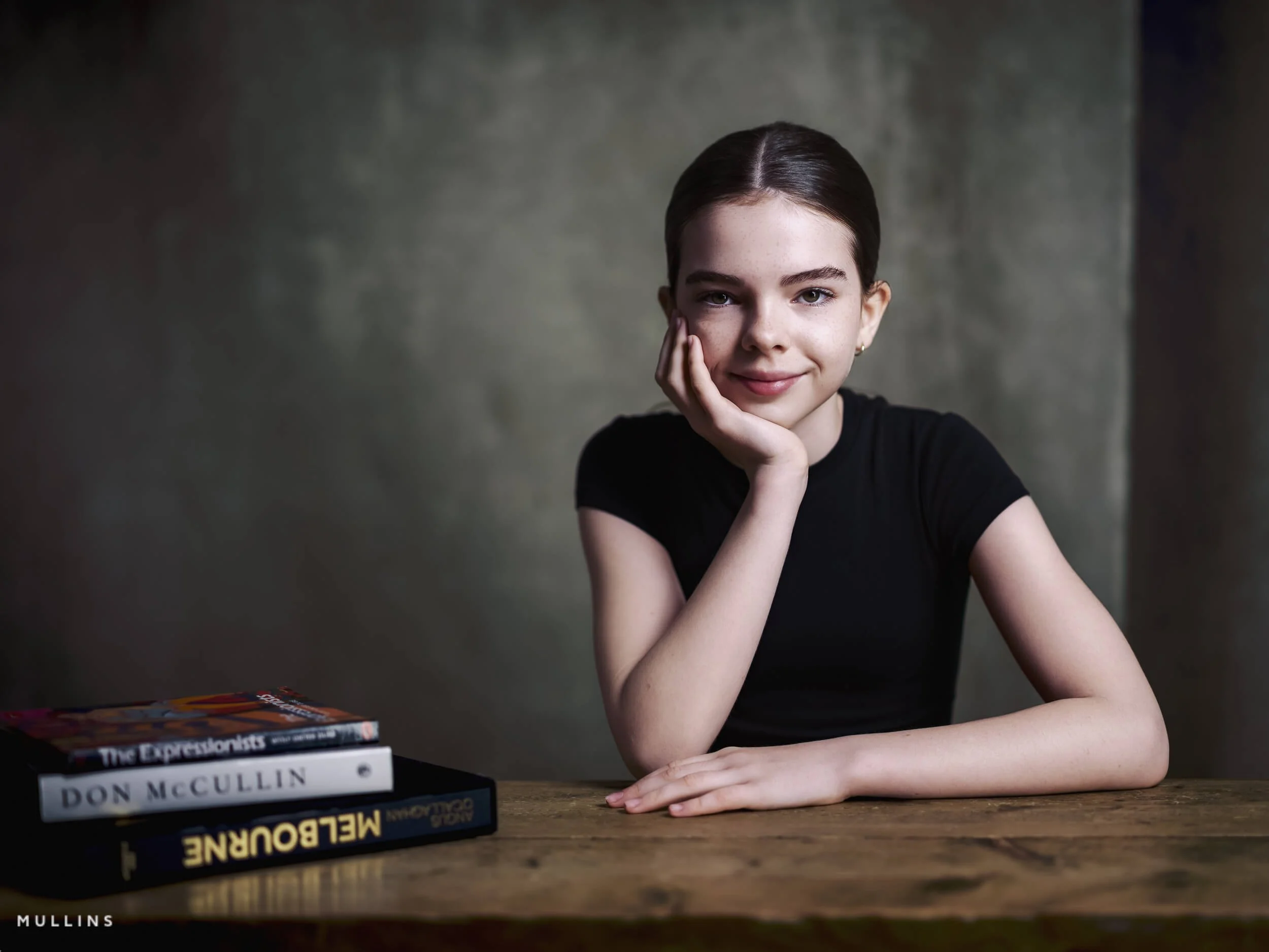 Portrait of a smiling young female actor seated at a wooden table with photography books beside her.