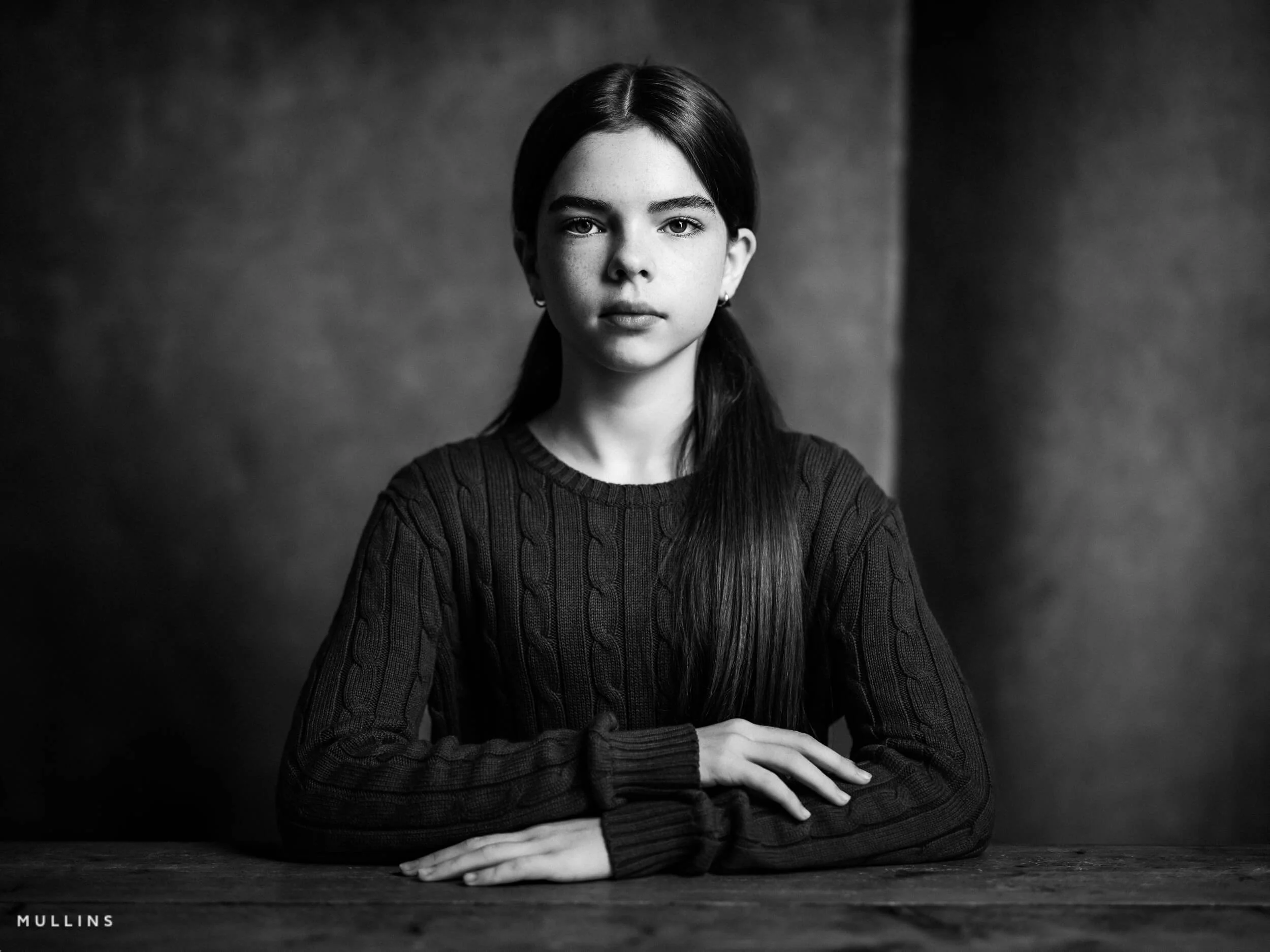 Black and white portrait of a young female actor seated at a wooden table with a calm expression, facing the camera.