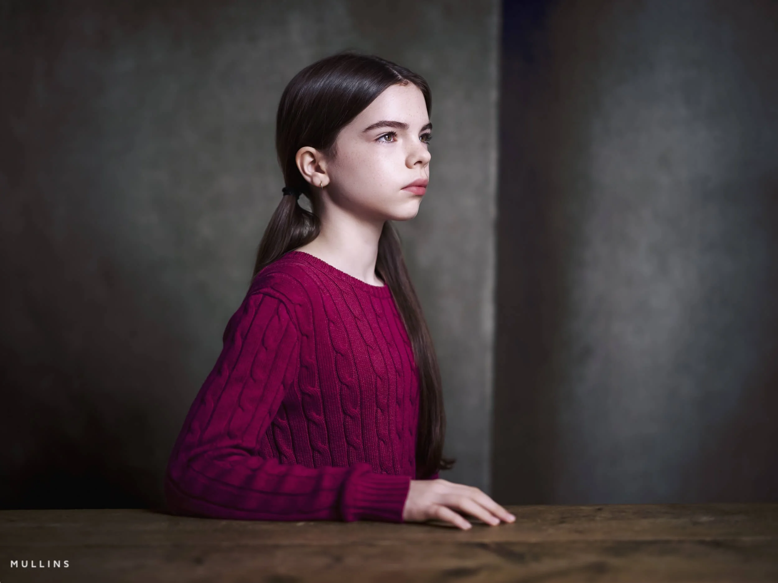 Side-profile portrait of a young female actor seated at a wooden table in the studio, wearing a bright jumper.
