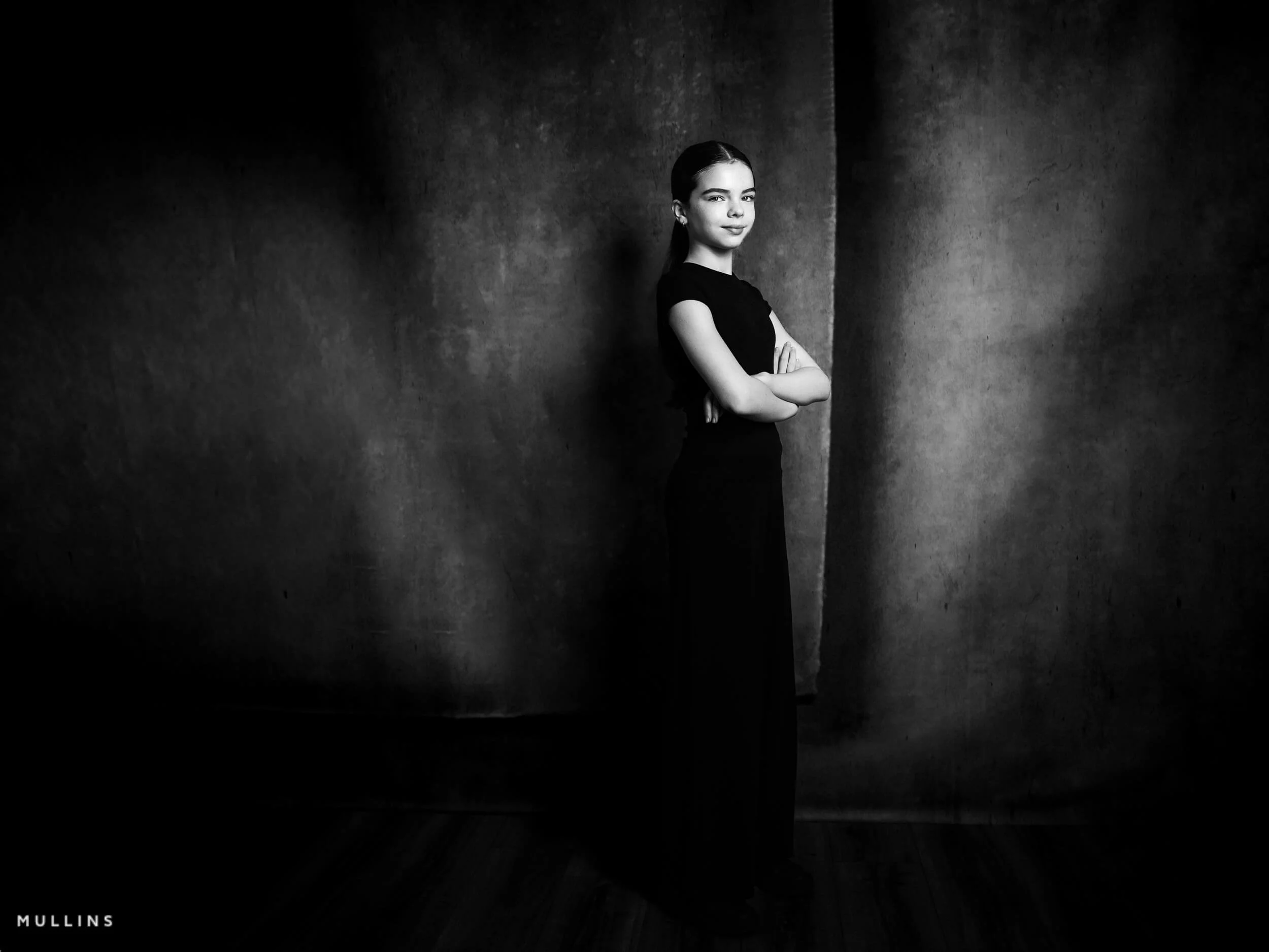 Black and white portrait of a young female actor standing in a black outfit against a textured studio backdrop.