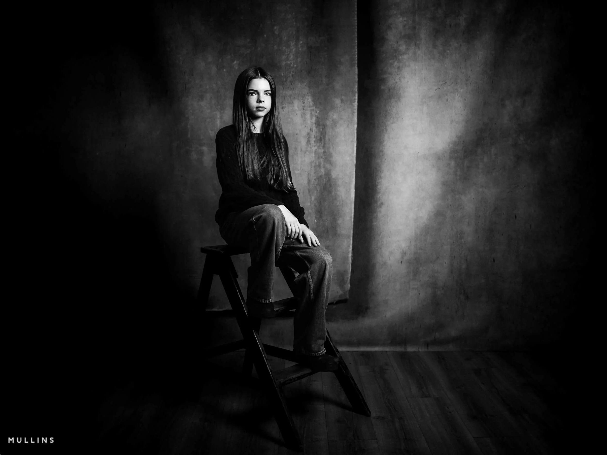 Black and white full-length studio portrait of a young female actor seated on a wooden step stool.