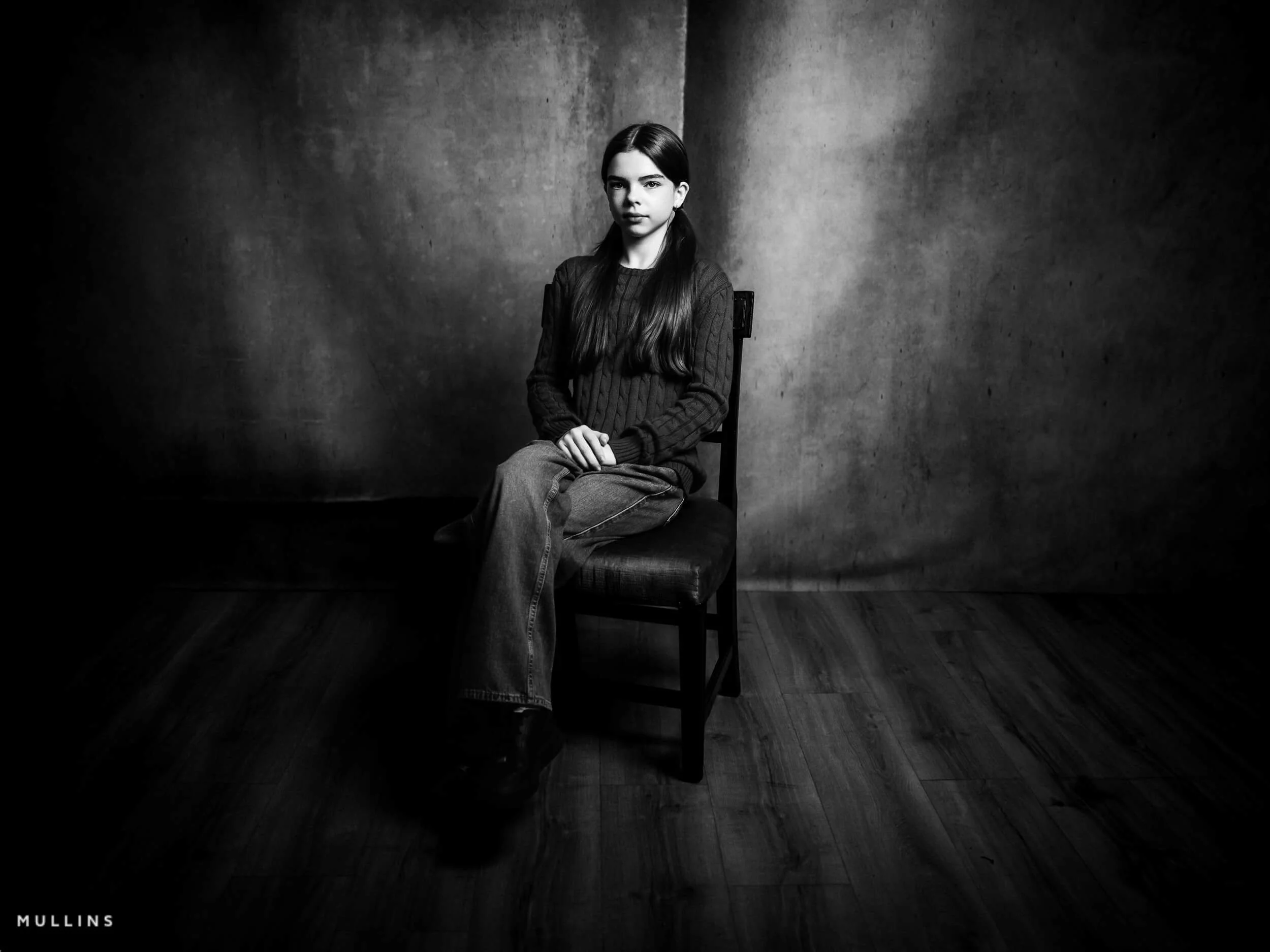 Black and white seated studio portrait of a young female actor wearing a jumper and jeans against a textured backdrop.