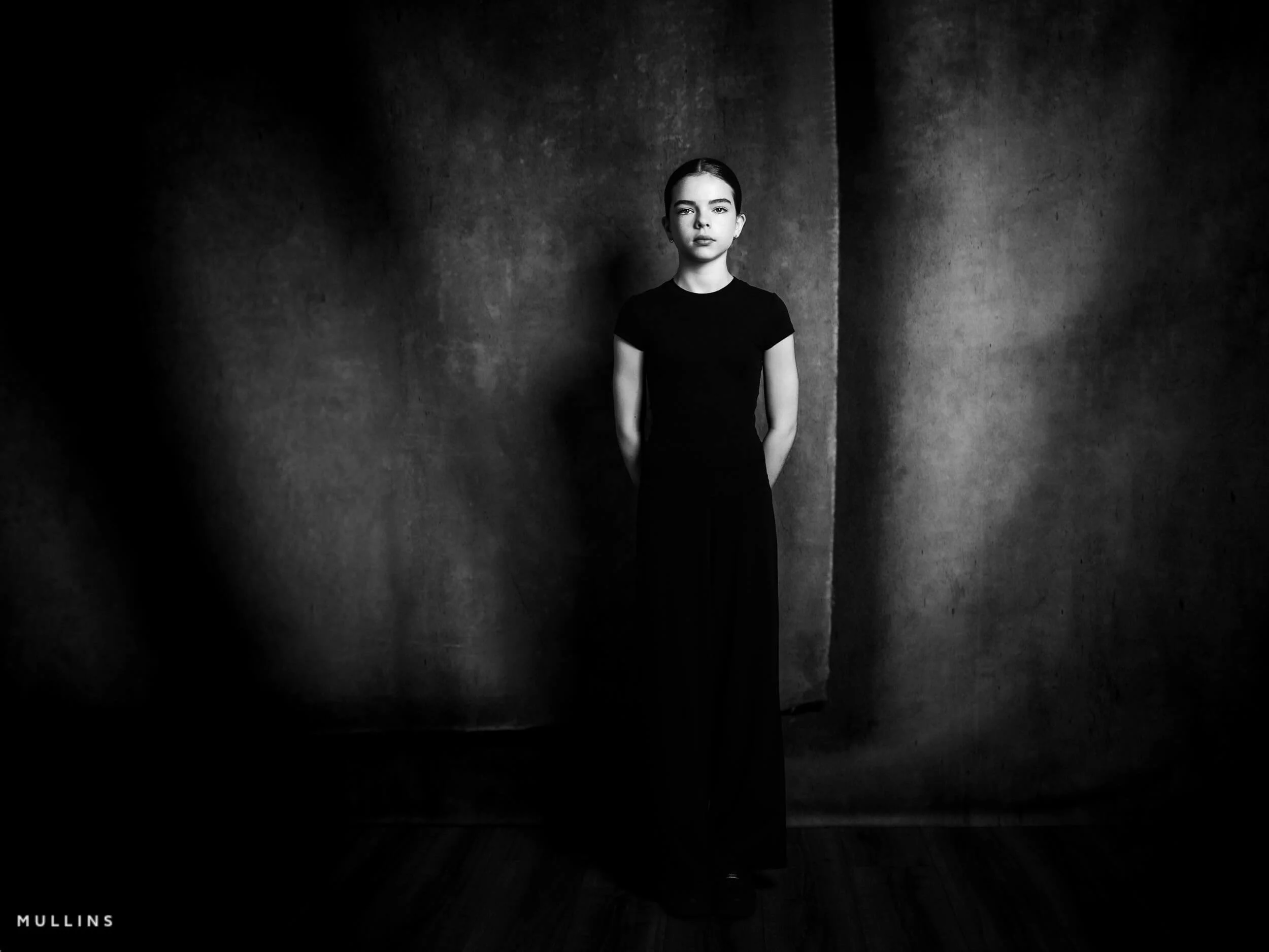 Black and white full-length portrait of a young female actor standing in a black outfit in the studio.
