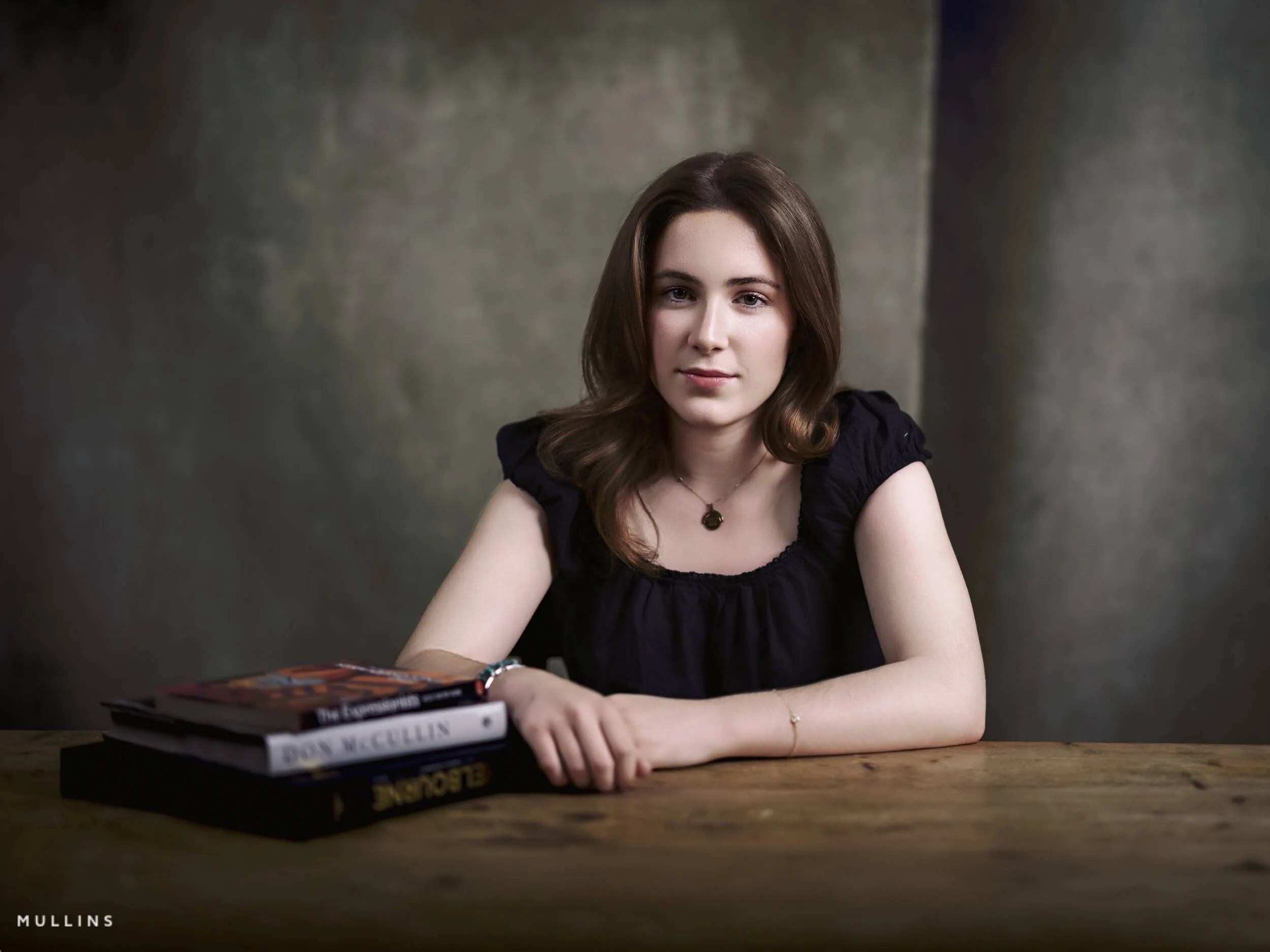 Actor portfolio photo of a young actress seated at a wooden table in a black top, looking directly at the camera.