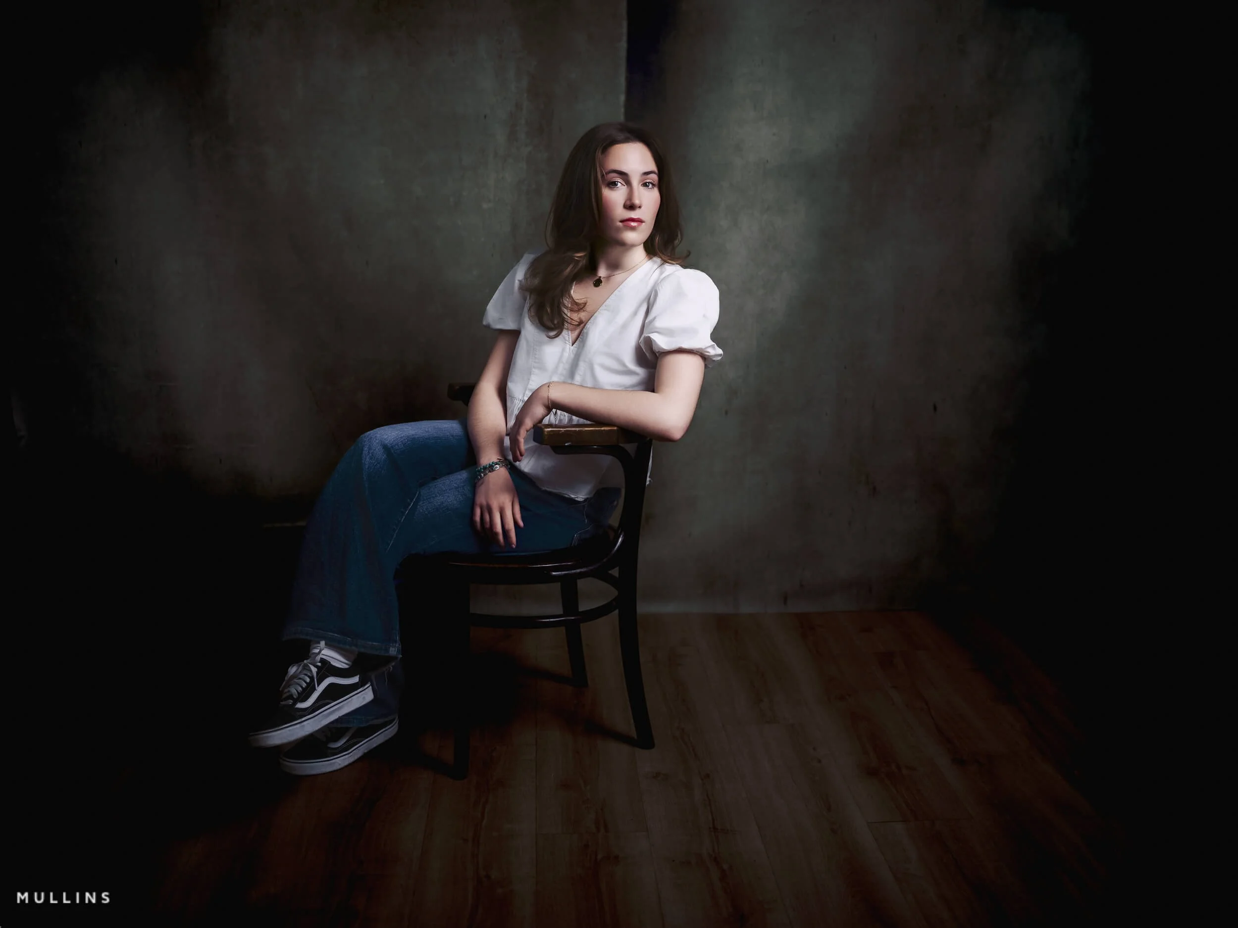 Actor portfolio photo of a young actress seated on a wooden chair in a white blouse and jeans, looking off to one side.
