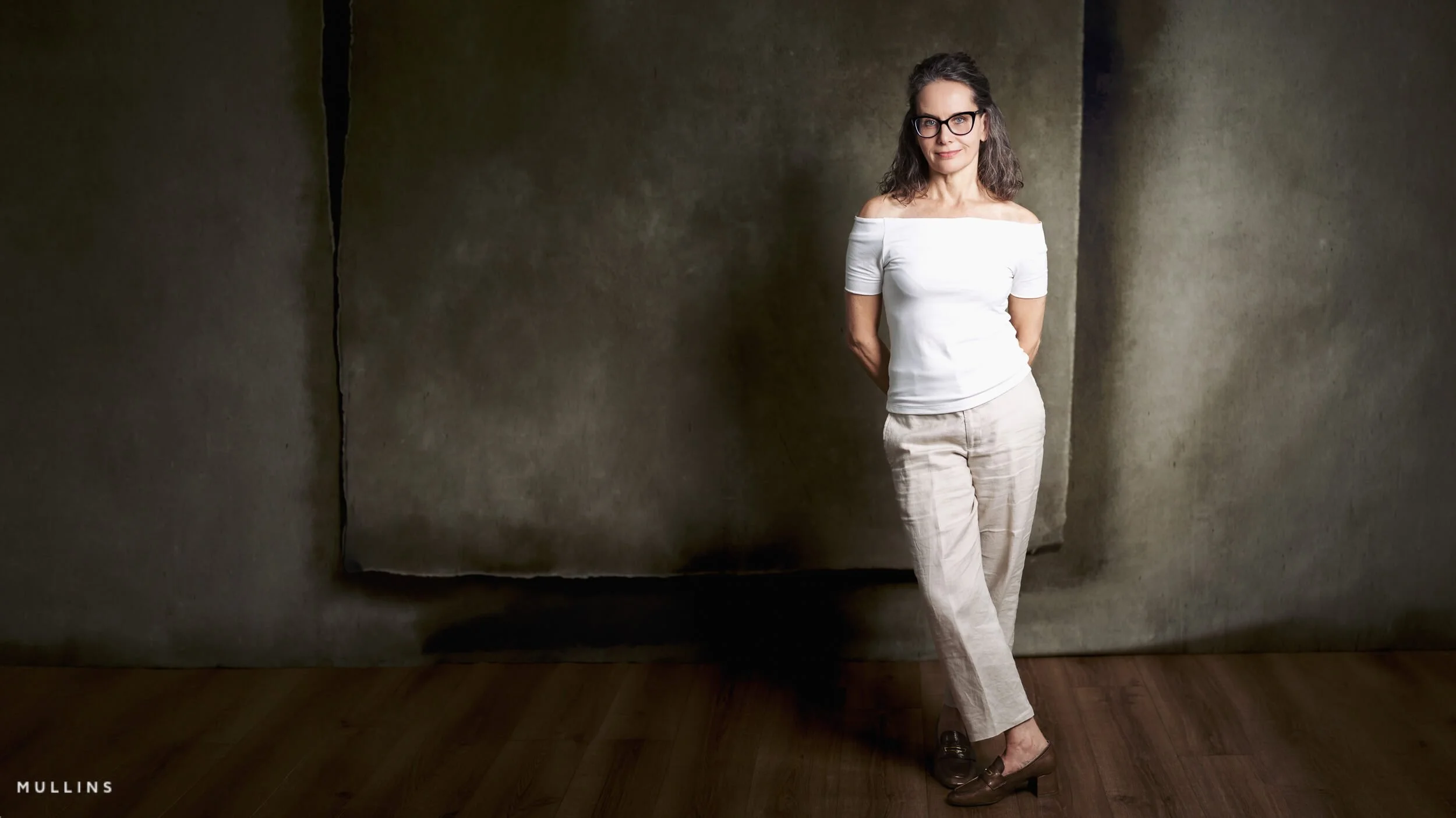 Studio portrait of a woman wearing glasses and light clothing, standing against a dark textured backdrop.