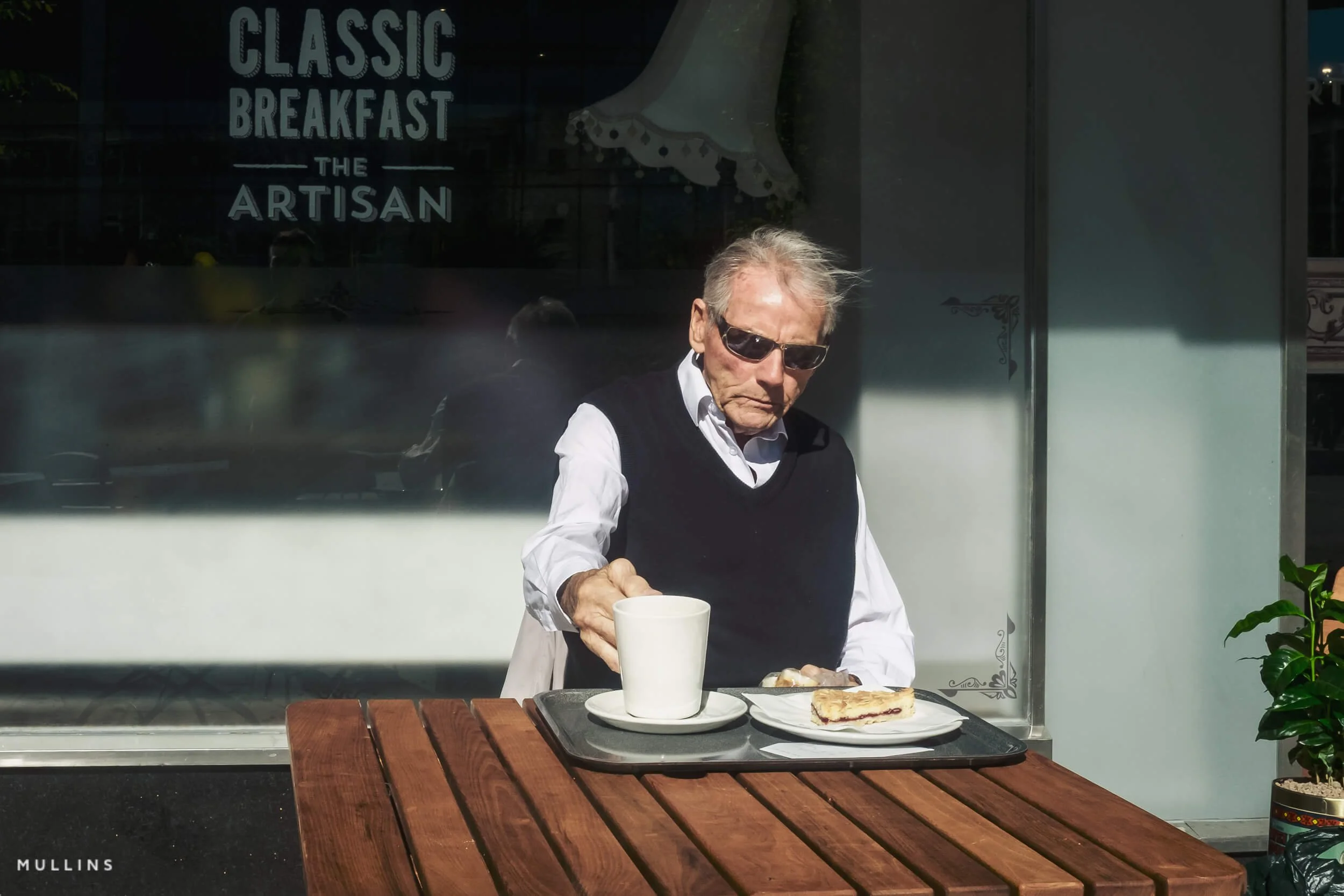 A man in sunglasses sitting outside a café with a cup and food on a tray.