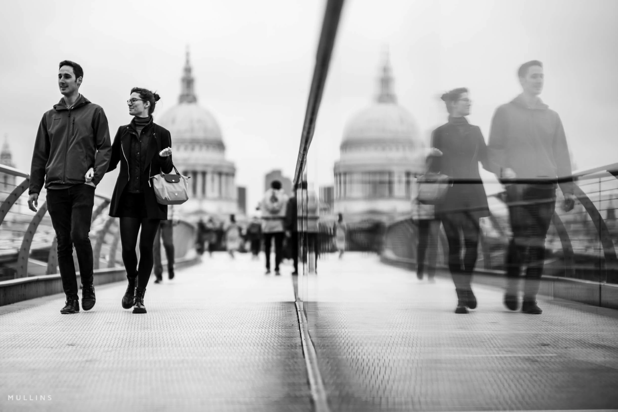 Two people walking across a pedestrian bridge beside a reflective glass barrier, with a domed building behind them.