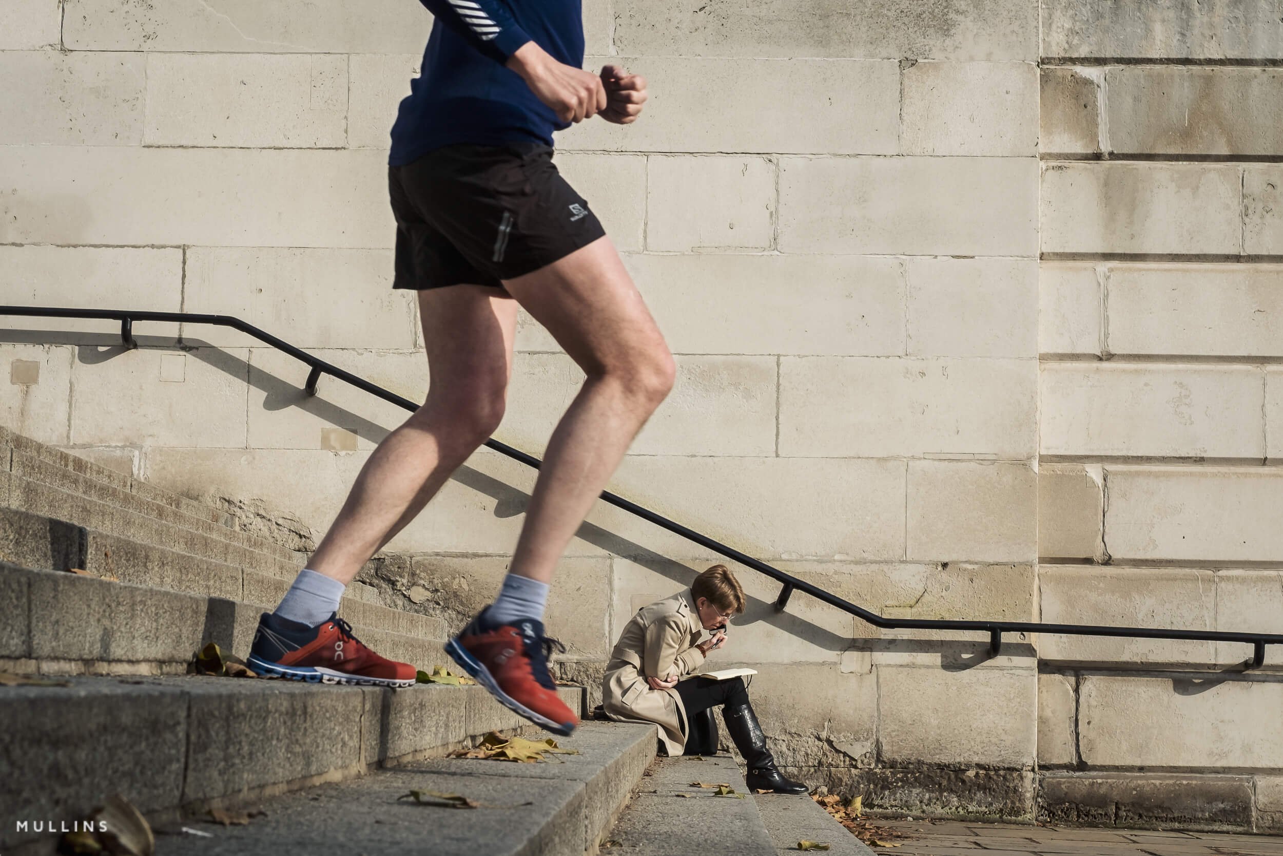 A runner’s legs cross the foreground on stone steps, with a person seated further down beside a pale wall.