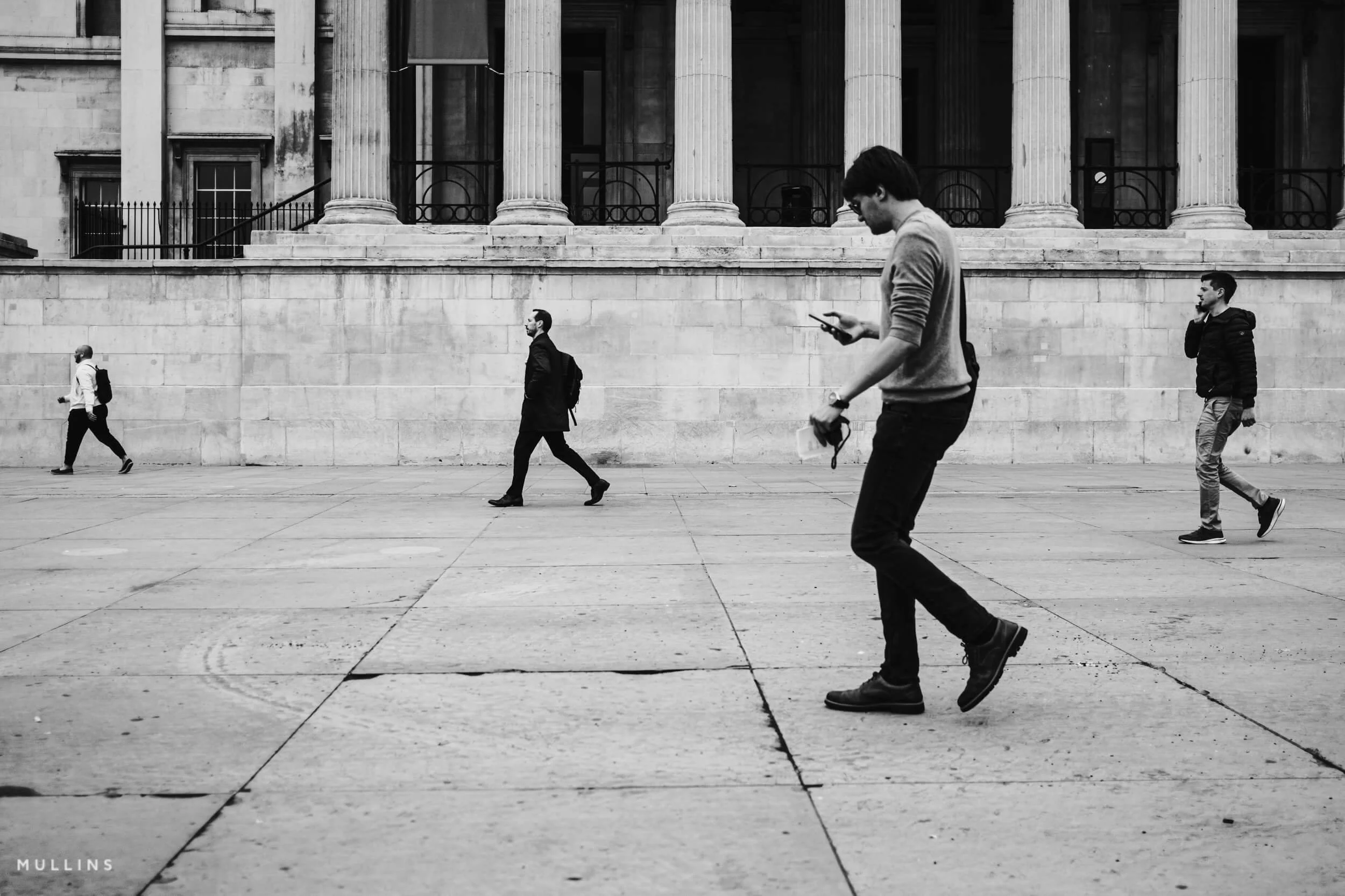 Four pedestrians spaced across a pavement in front of a columned stone building.