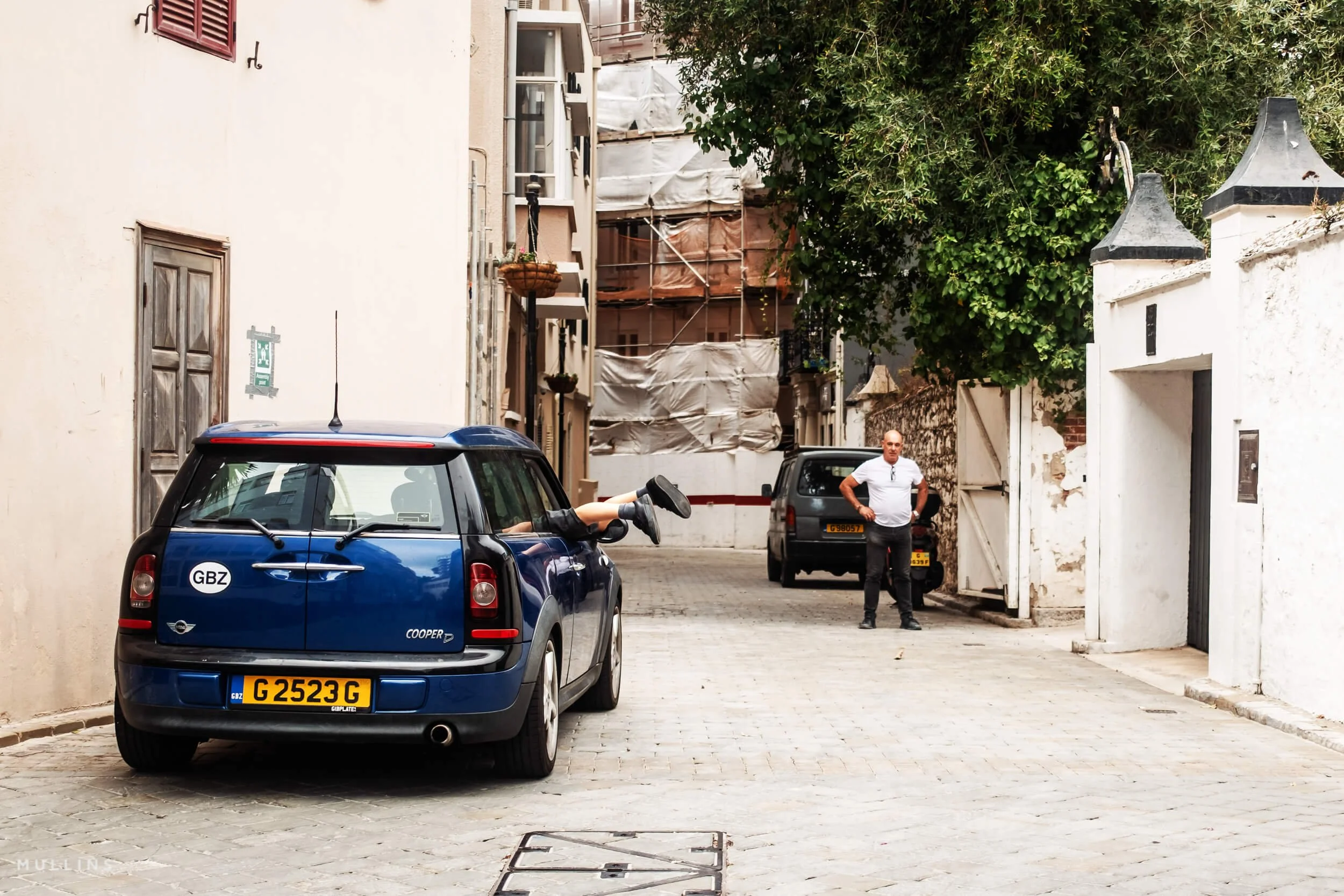 Legs extending from the side window of a blue Mini, with a man standing further along a narrow lane.