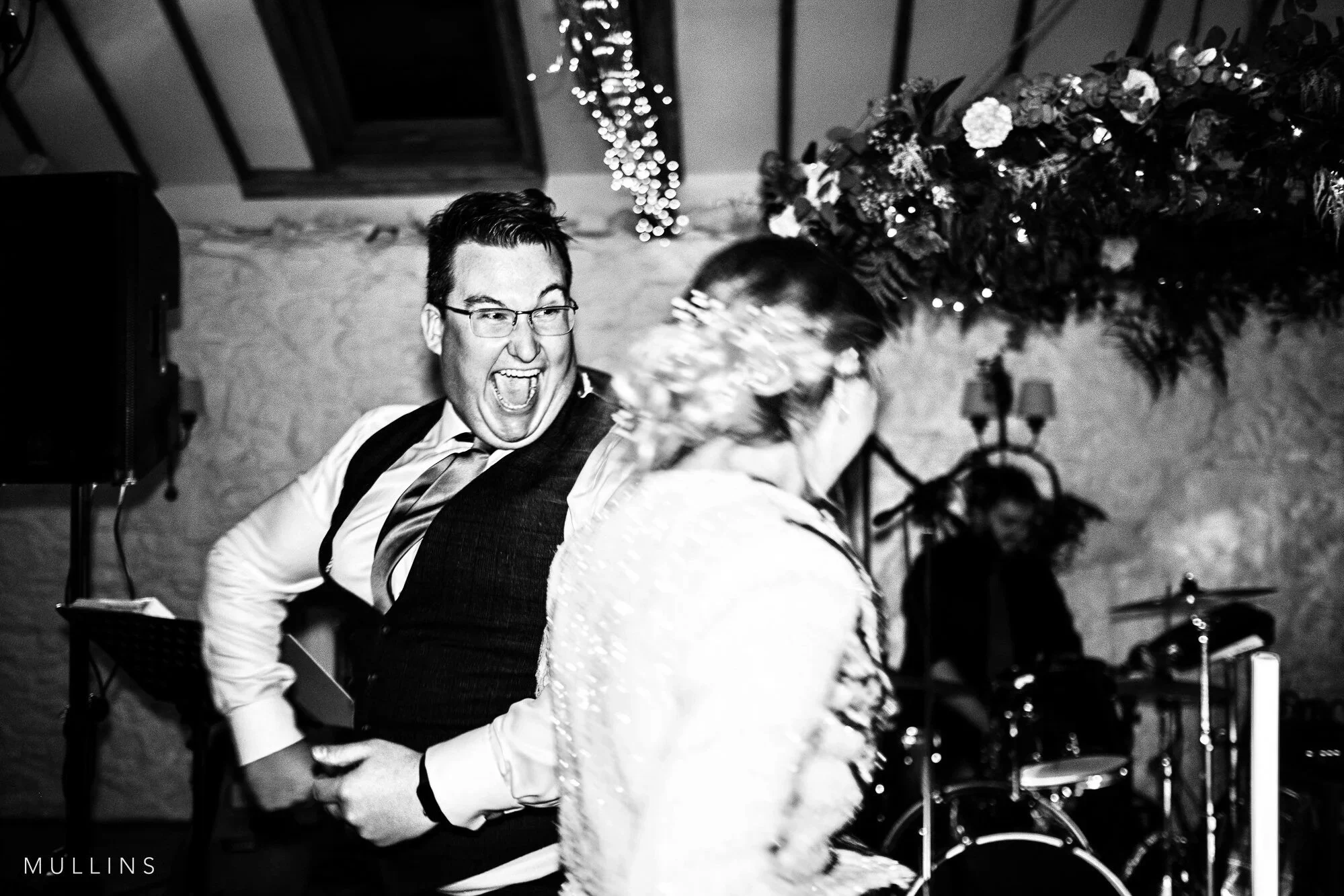 Two people dancing at a celebration event, one man wearing glasses and a vest, and a woman in a lace dress with a floral headpiece, in a room decorated with string lights and flowers.