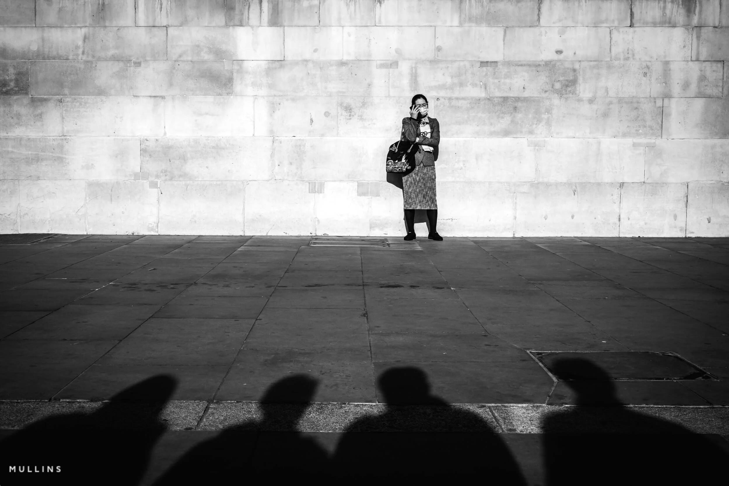 A person standing against a pale stone wall, with four people’s shadows across the foreground.