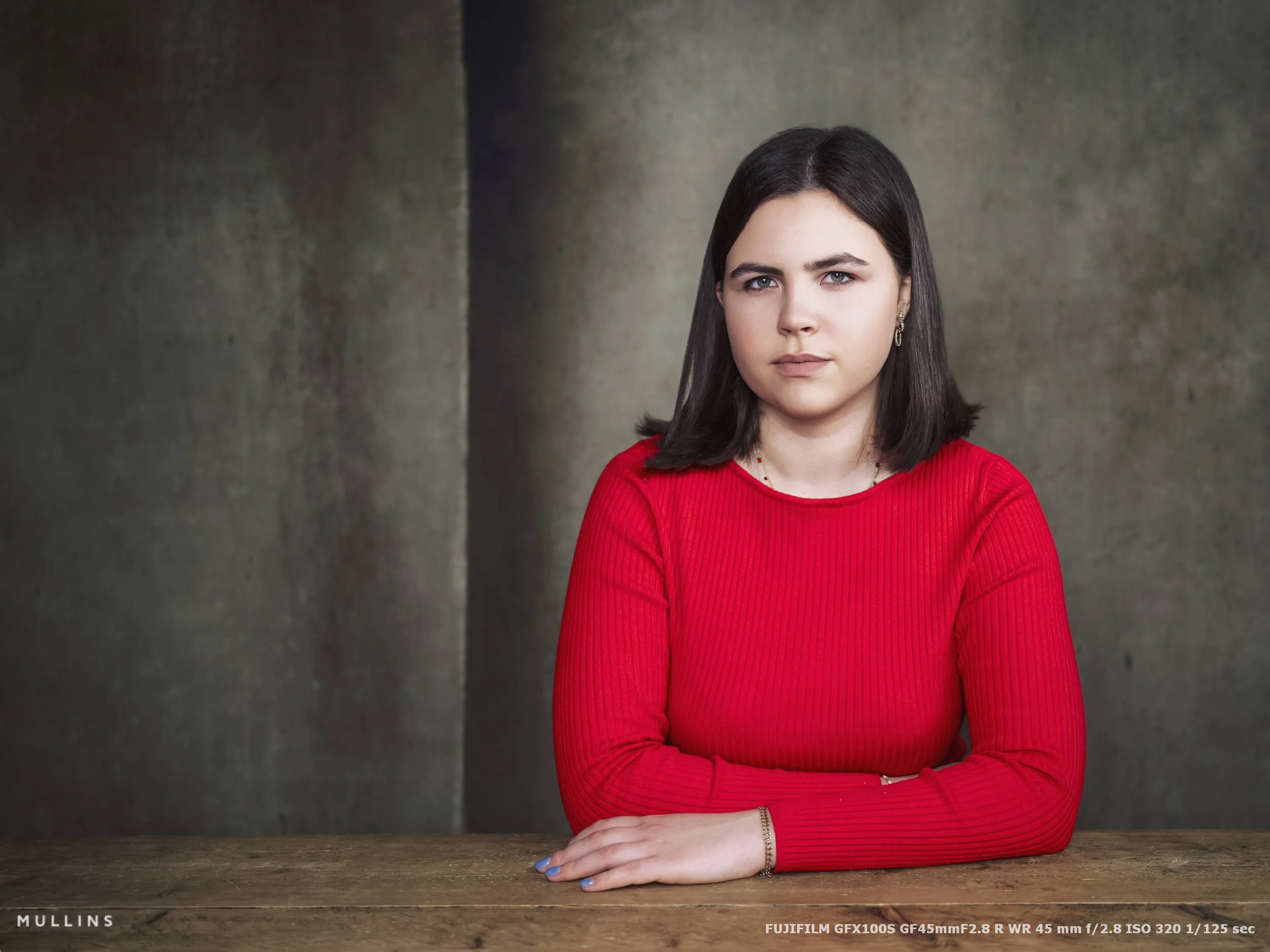 A girl seated at a wooden table with arms folded lightly in front of her, photographed in soft studio light with the GF45mm lens.