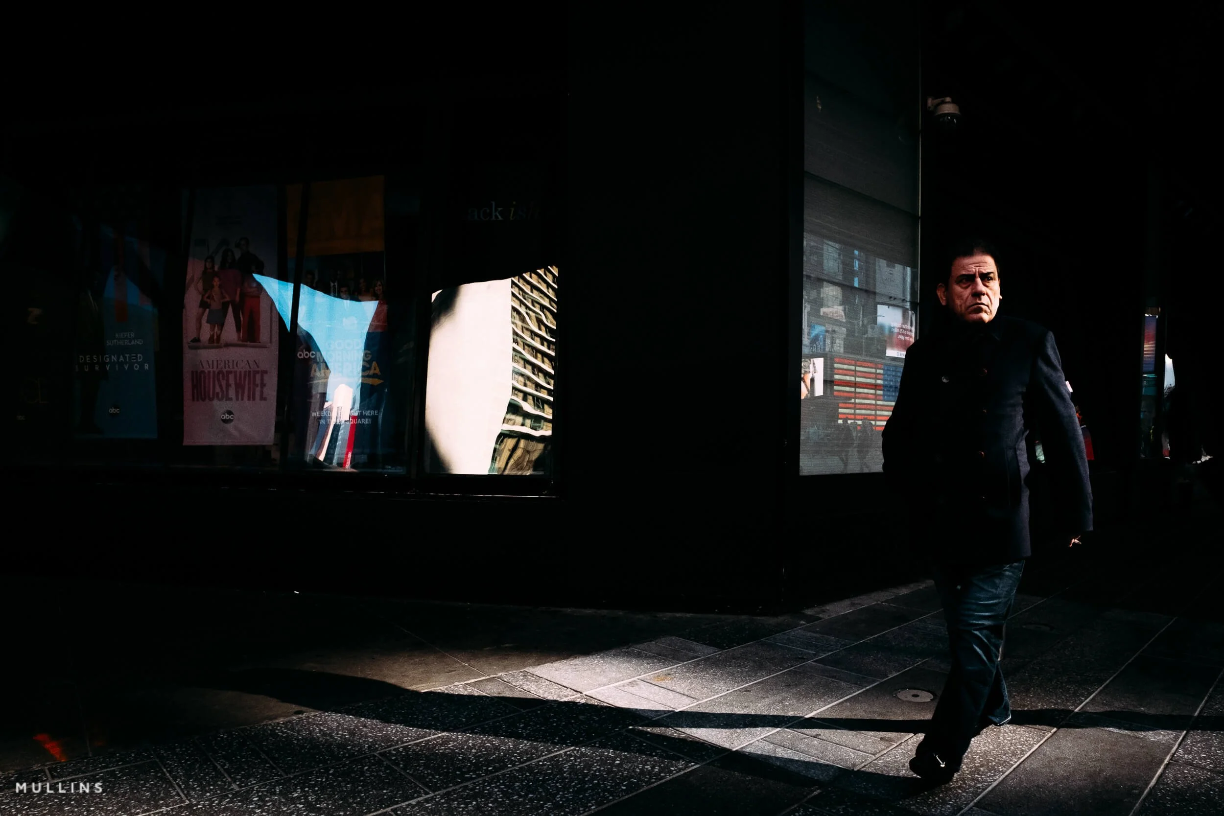 A man walking past dark windows, with light catching his face, reflections in the glass and a long shadow across the pavement.