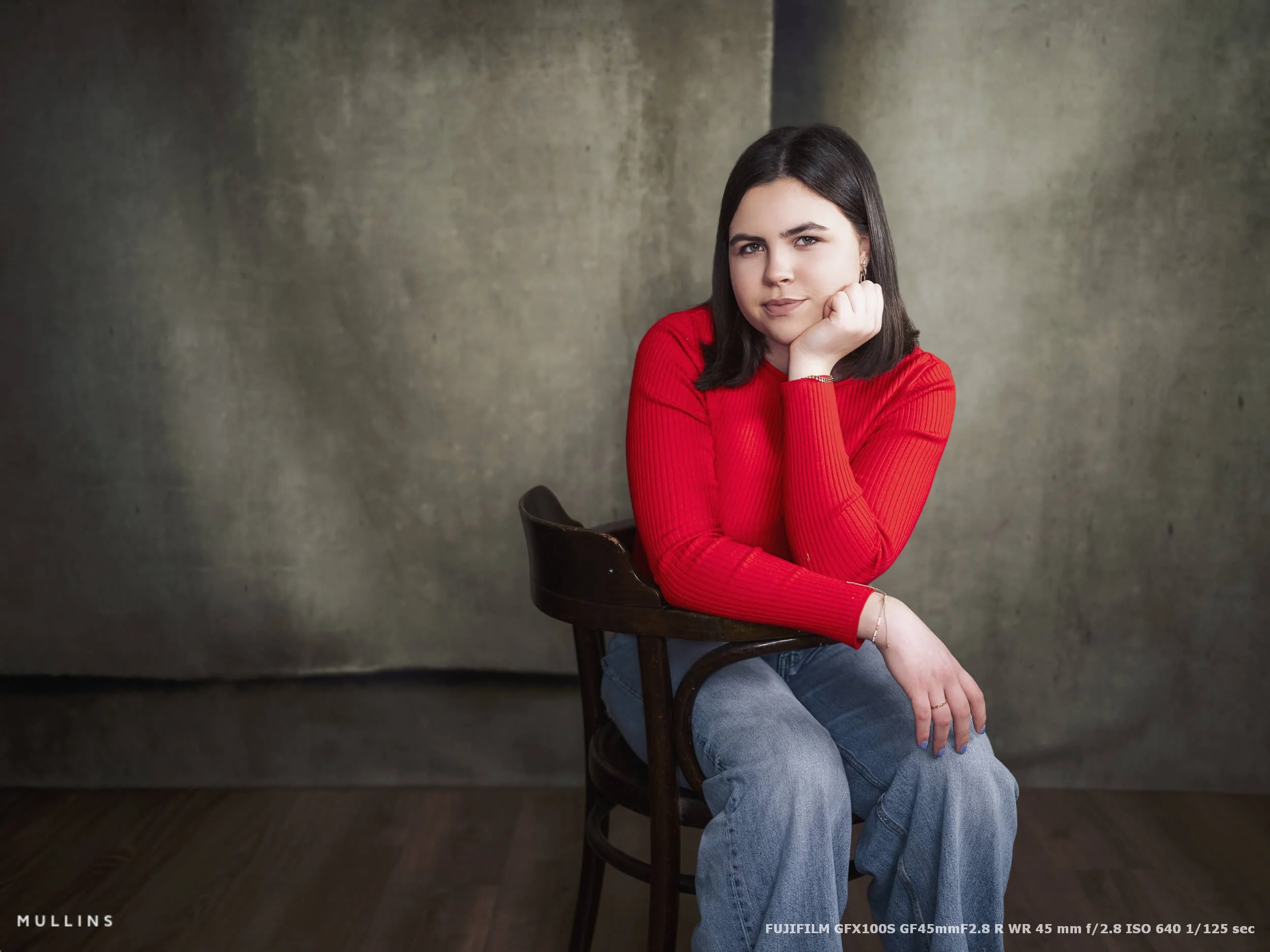 Girl sitting sideways on a wooden chair in the studio with her chin resting on her hand, photographed with a wider environmental composition on the GF45mm lens.