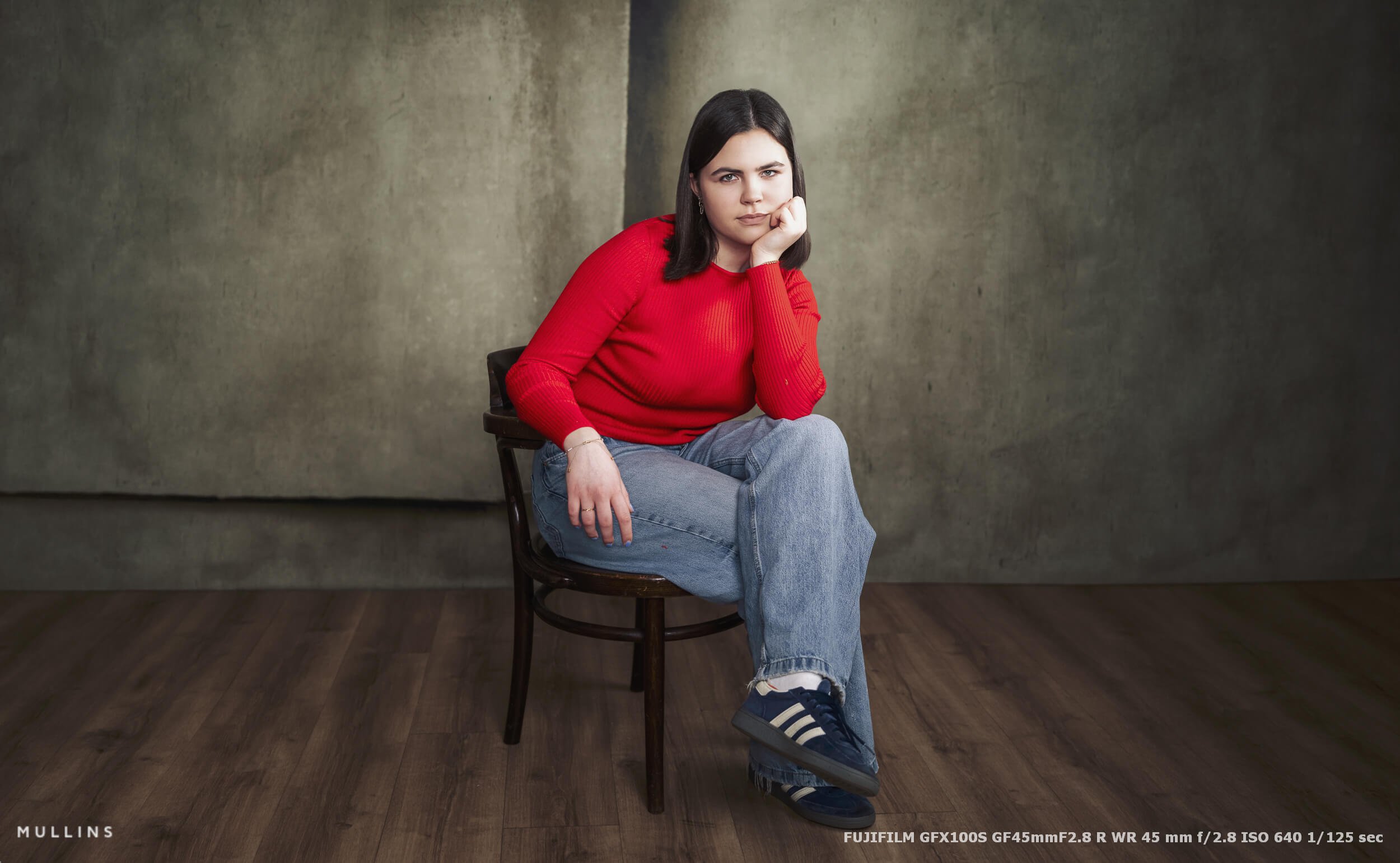 Girl leaning forward slightly while seated on a wooden chair, framed with negative space and studio floor visible, photographed on Fujifilm GFX100S with GF45mm lens.