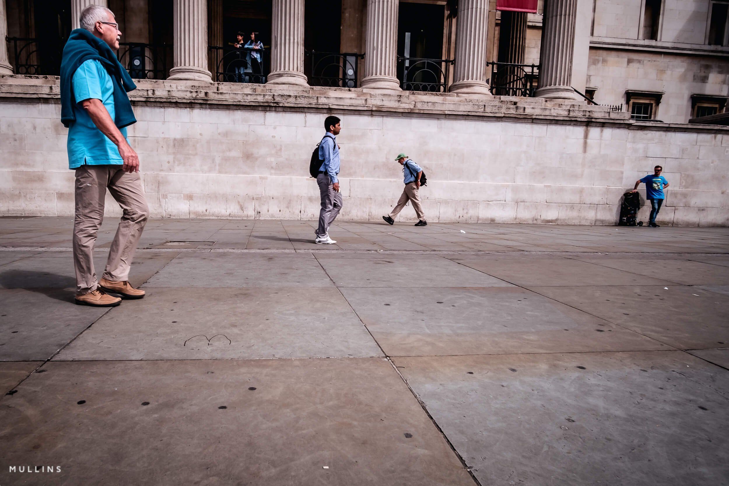 People wearing different shades of blue, spaced across a pavement beside a pale stone wall and columns.