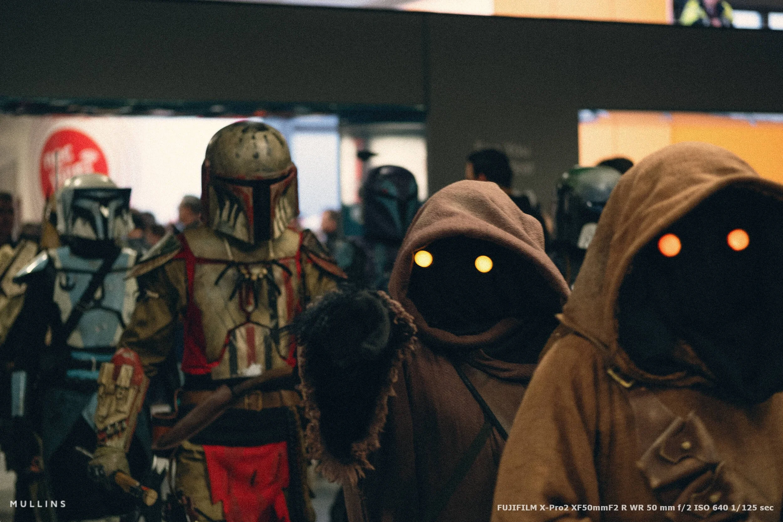 Two hooded sci-fi cosplayers with glowing eyes in the foreground, with armoured costumed figures behind them at a convention.