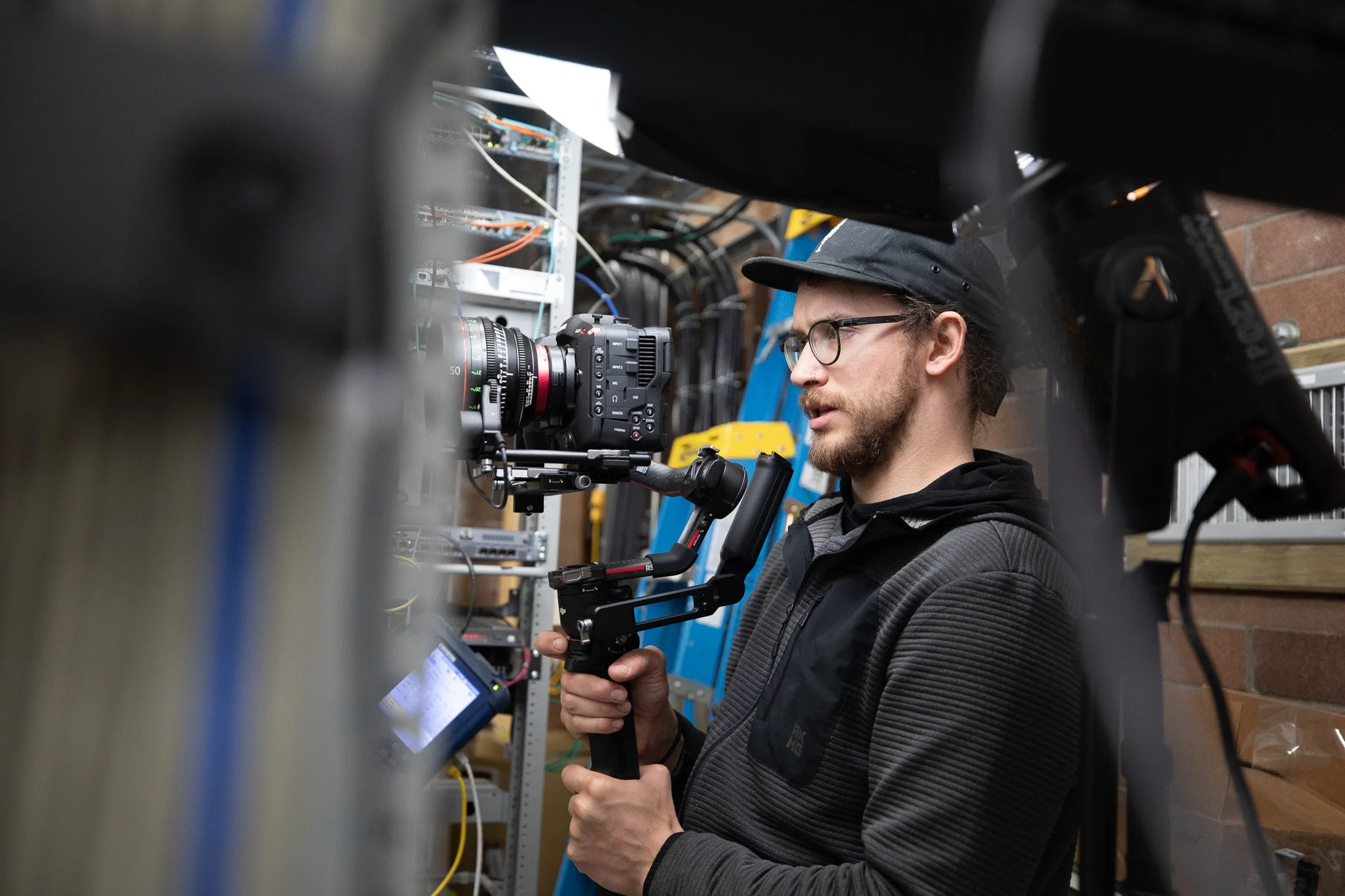 A man operating a professional camera mounted on a stabilizer in a tech or wiring room with shelves of electronic equipment.