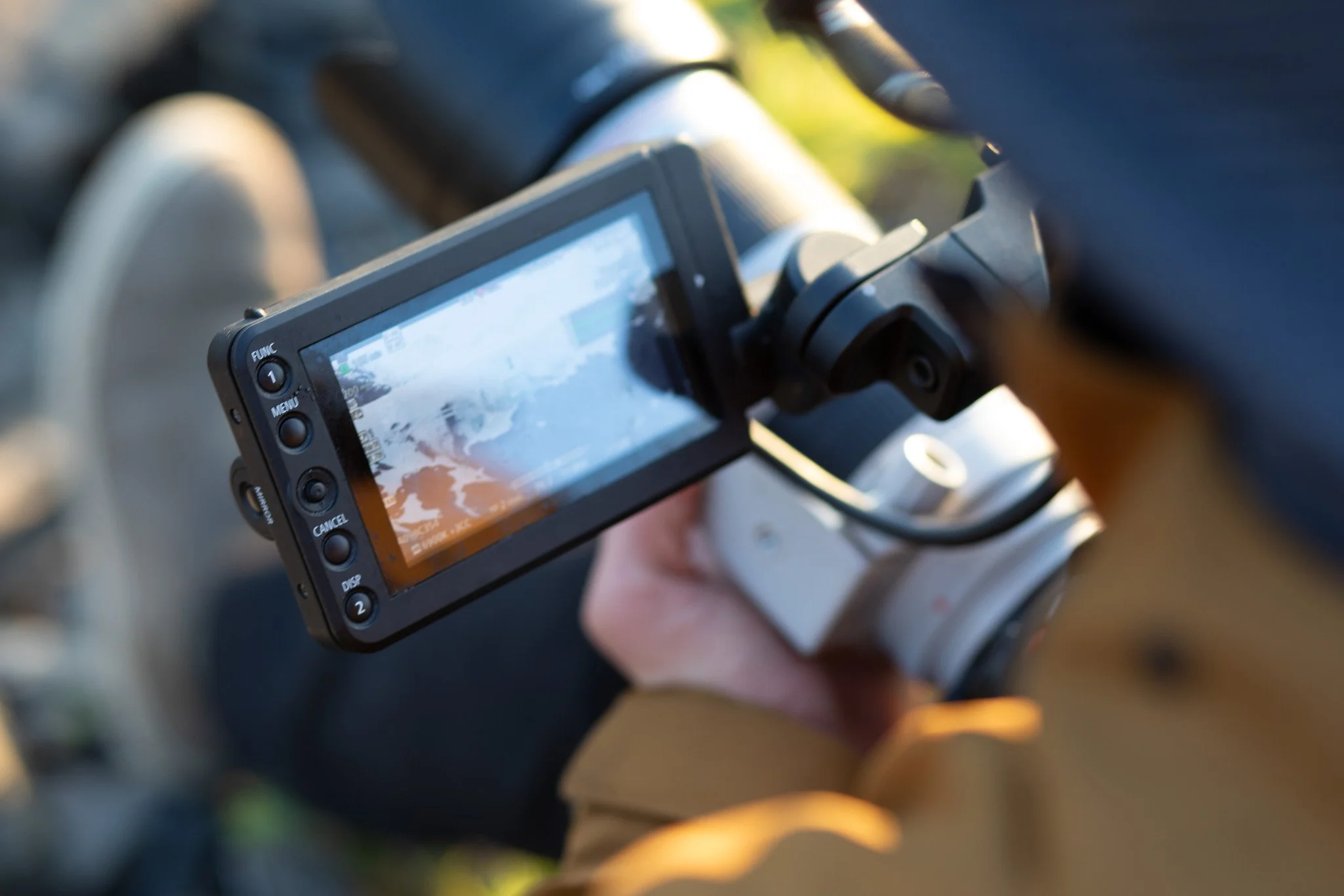 Person holding a handheld camera with a screen displaying a viewfinder, capturing an outdoor scene.