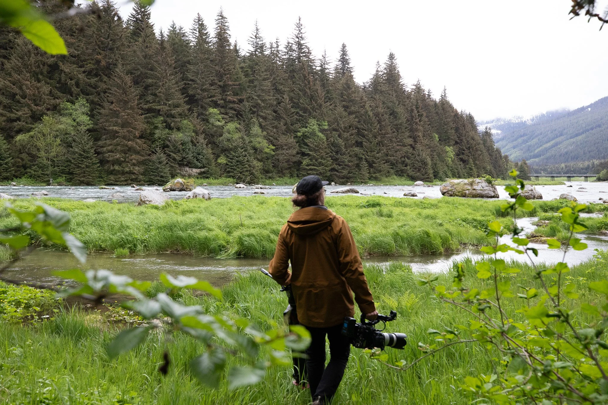 Person holding a camera and walking near a river in a lush green forested area with mountains and a cloudy sky in the background.