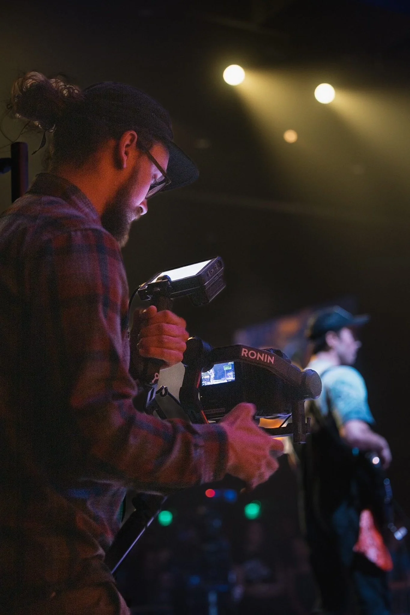 A videographer with a beard, glasses, and a cap is filming a performer on stage using a professional camera, with stage lights illuminating the scene.
