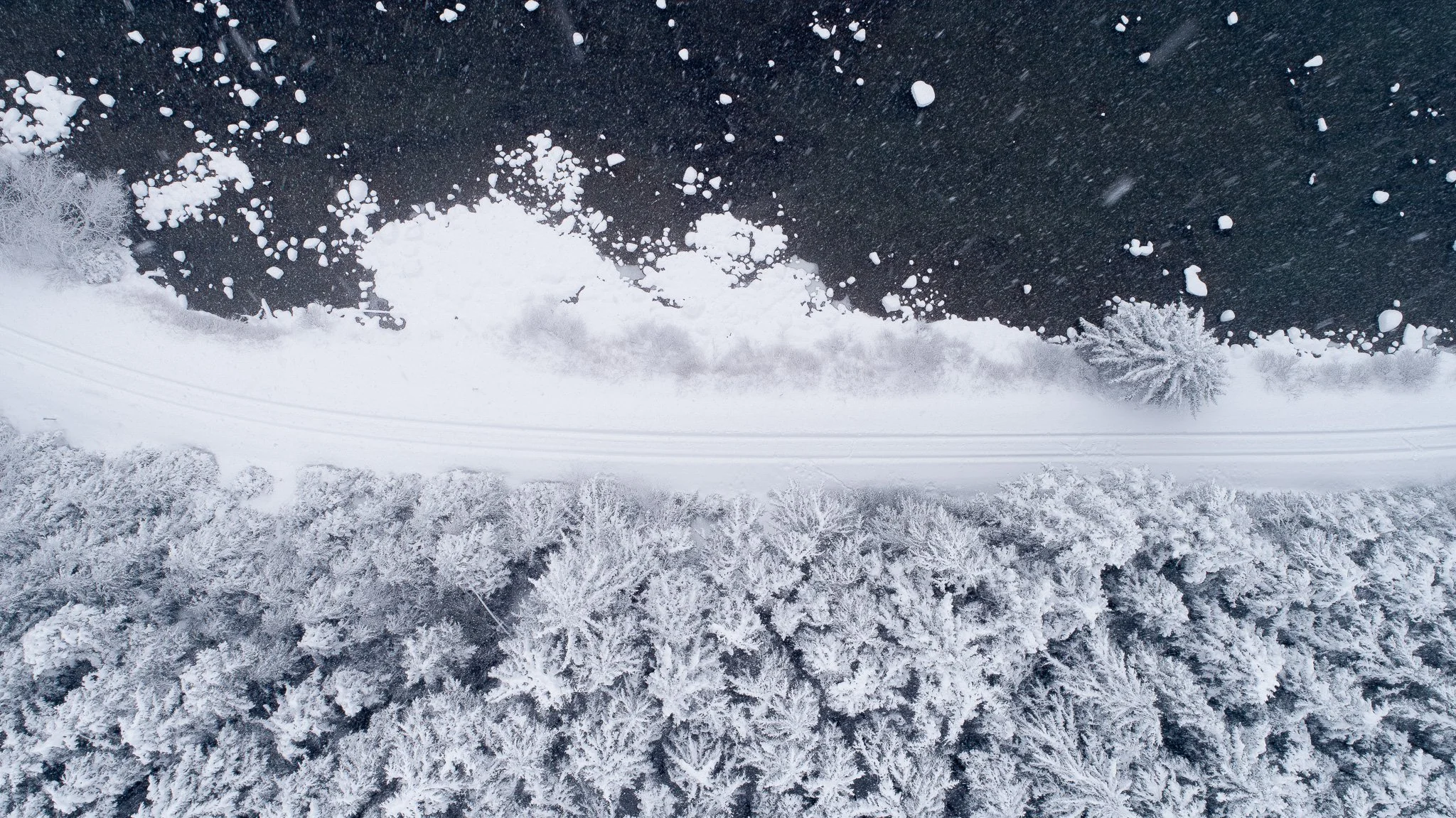 Aerial view of a snow-covered forest with a snow-covered road running through it, frozen lake with ice and snow patches, and snow-topped trees.