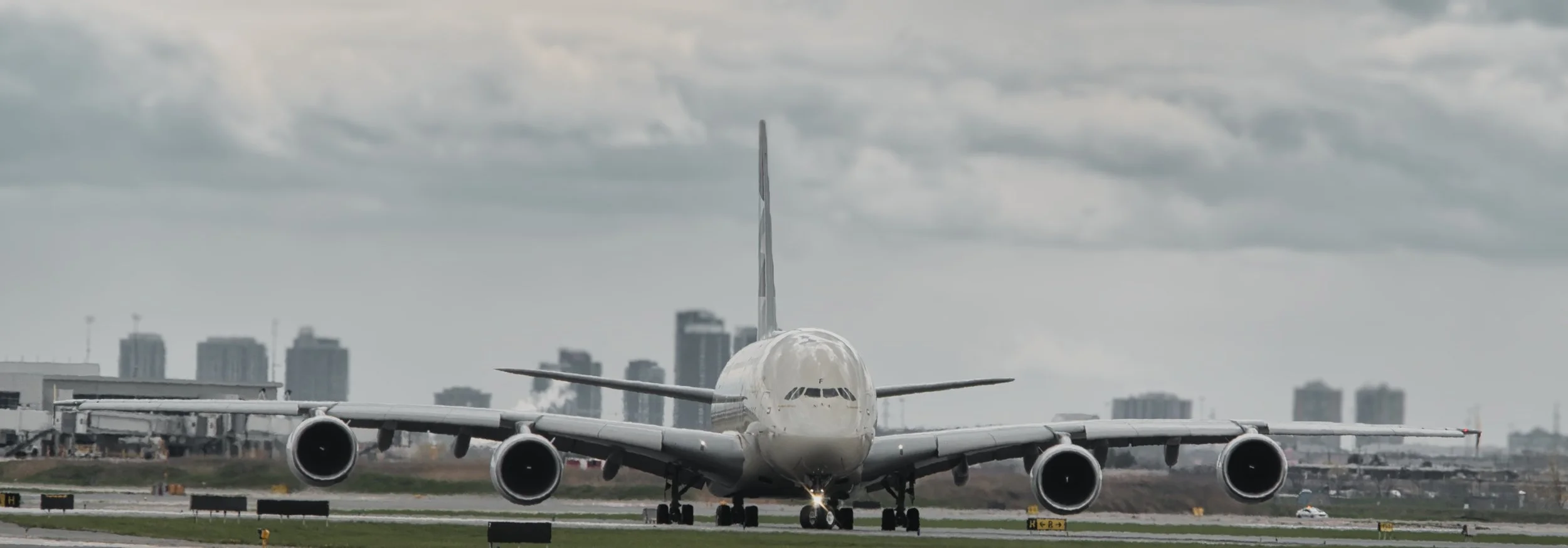 A large white commercial airplane on the runway, facing towards the camera, with city buildings and cloudy sky in the background.