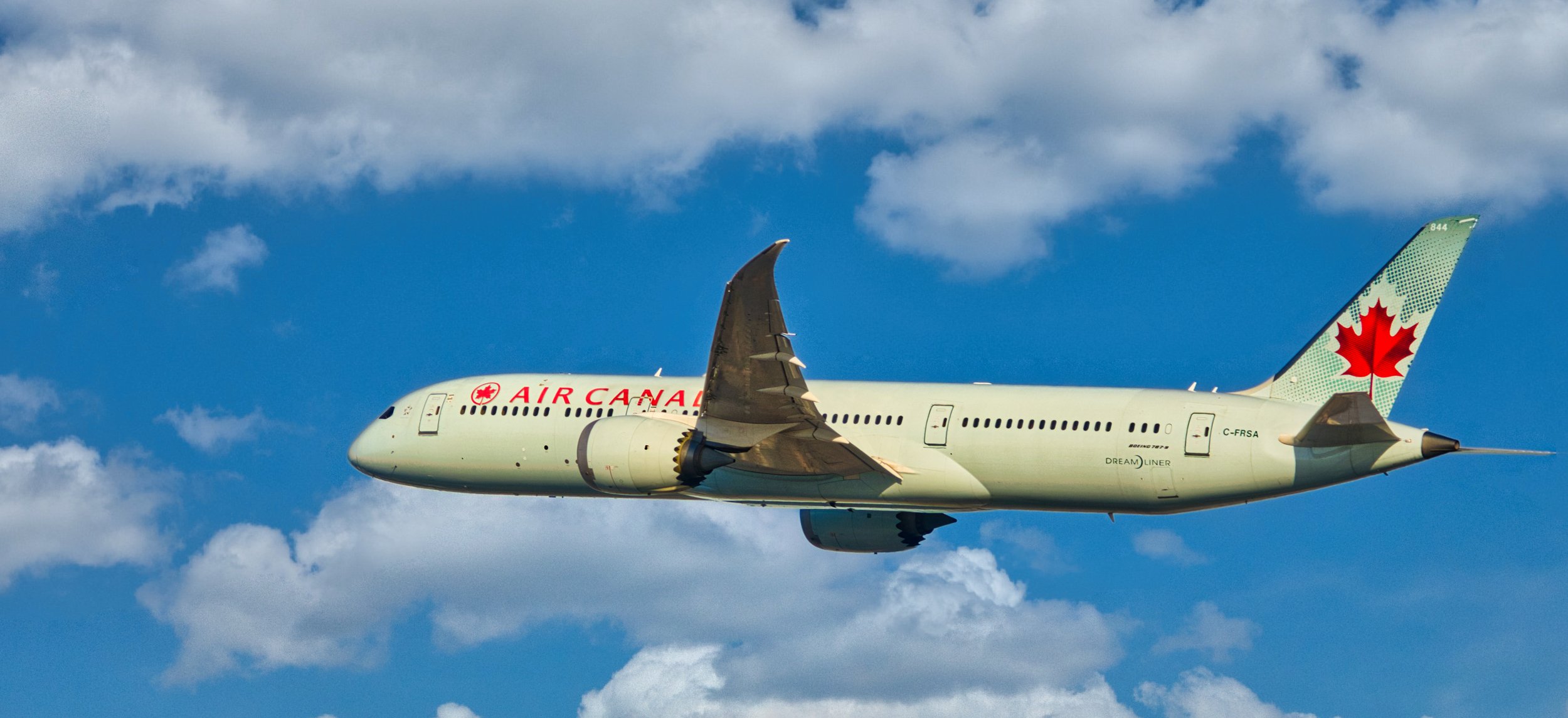An Air Canada airplane flying in a blue sky with scattered clouds, featuring a Canadian flag on its tail and the words 'Air Canada' on the fuselage.