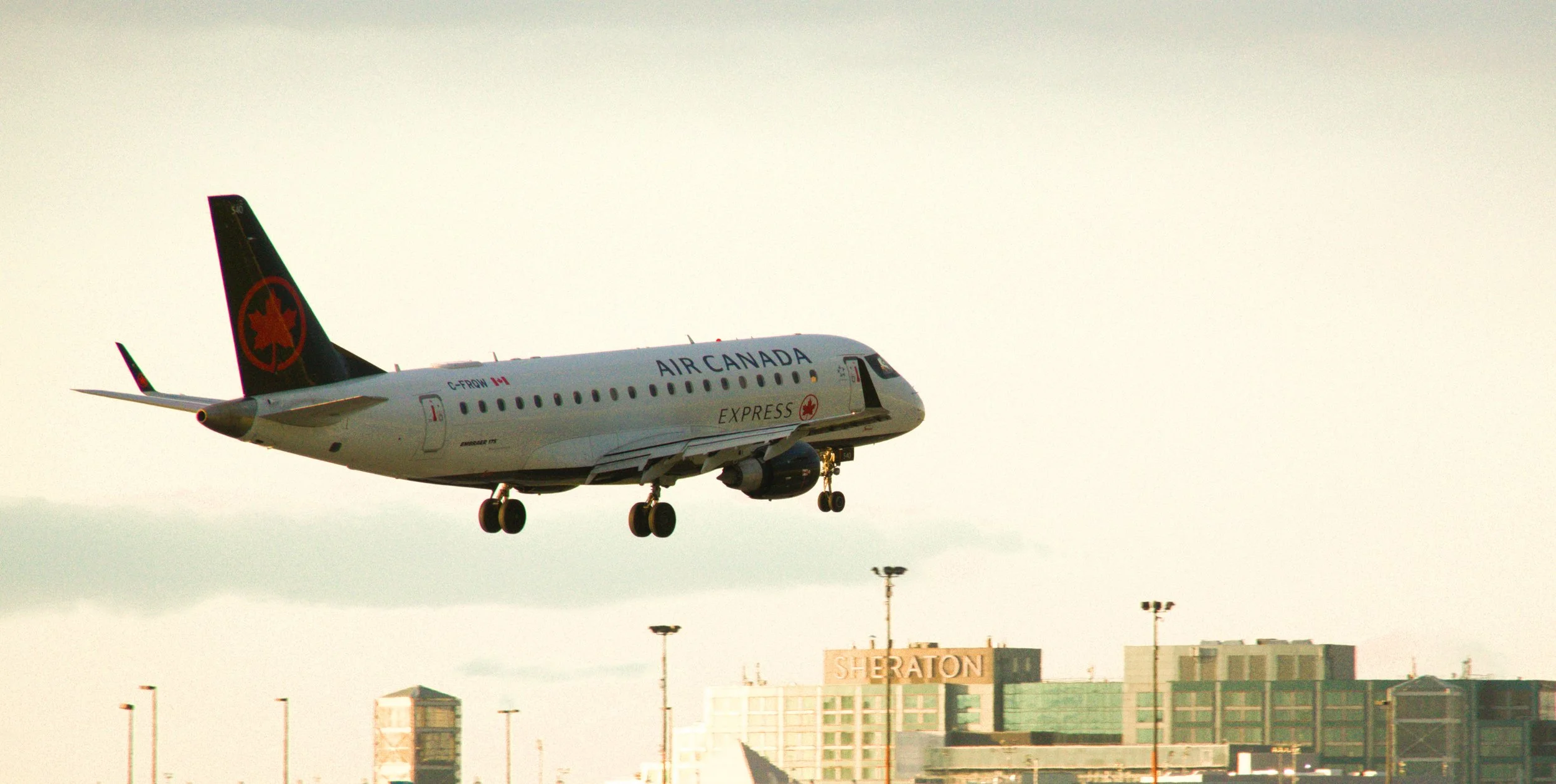 An Air Canada Express airplane flying low, with the Sheraton hotel in the background and lampposts below.