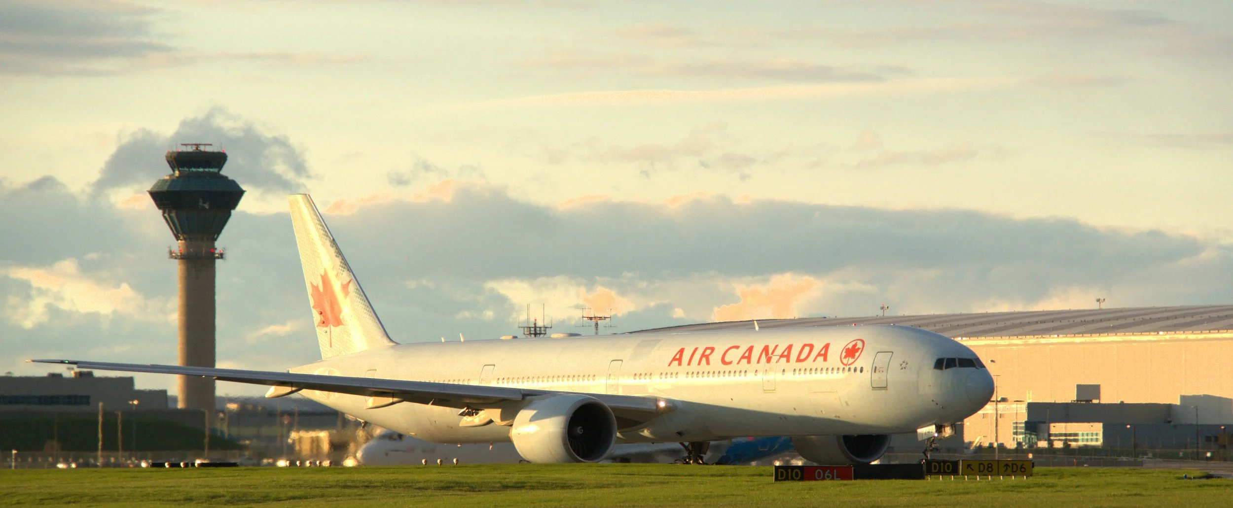 Air Canada airplane on the runway at an airport with an air traffic control tower in the background under a sky with scattered clouds.