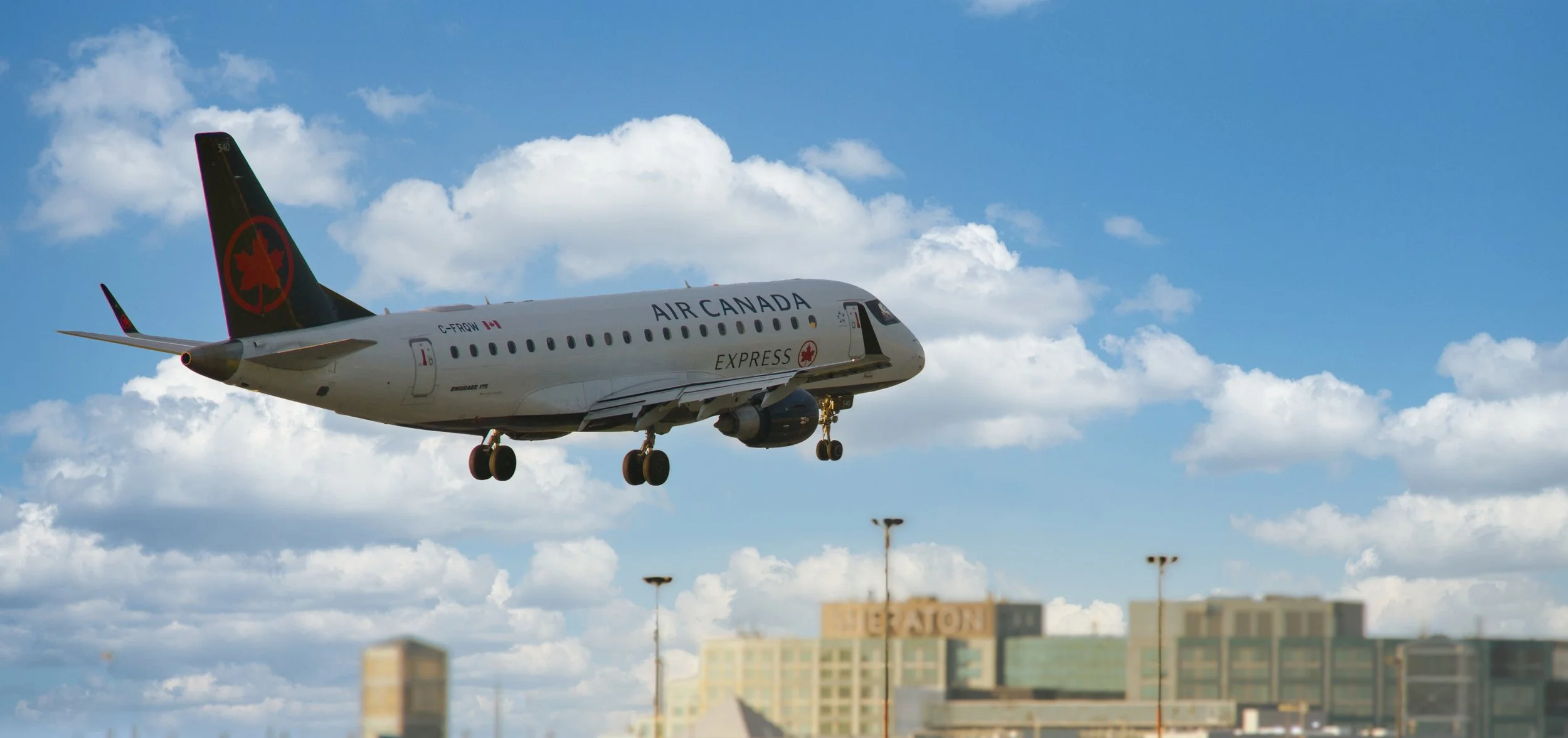 An Air Canada Express airplane is flying in the sky with a backdrop of white clouds and blue sky. The airplane is approaching a landing, with its landing gear extended. In the background, there are buildings and streetlights below.