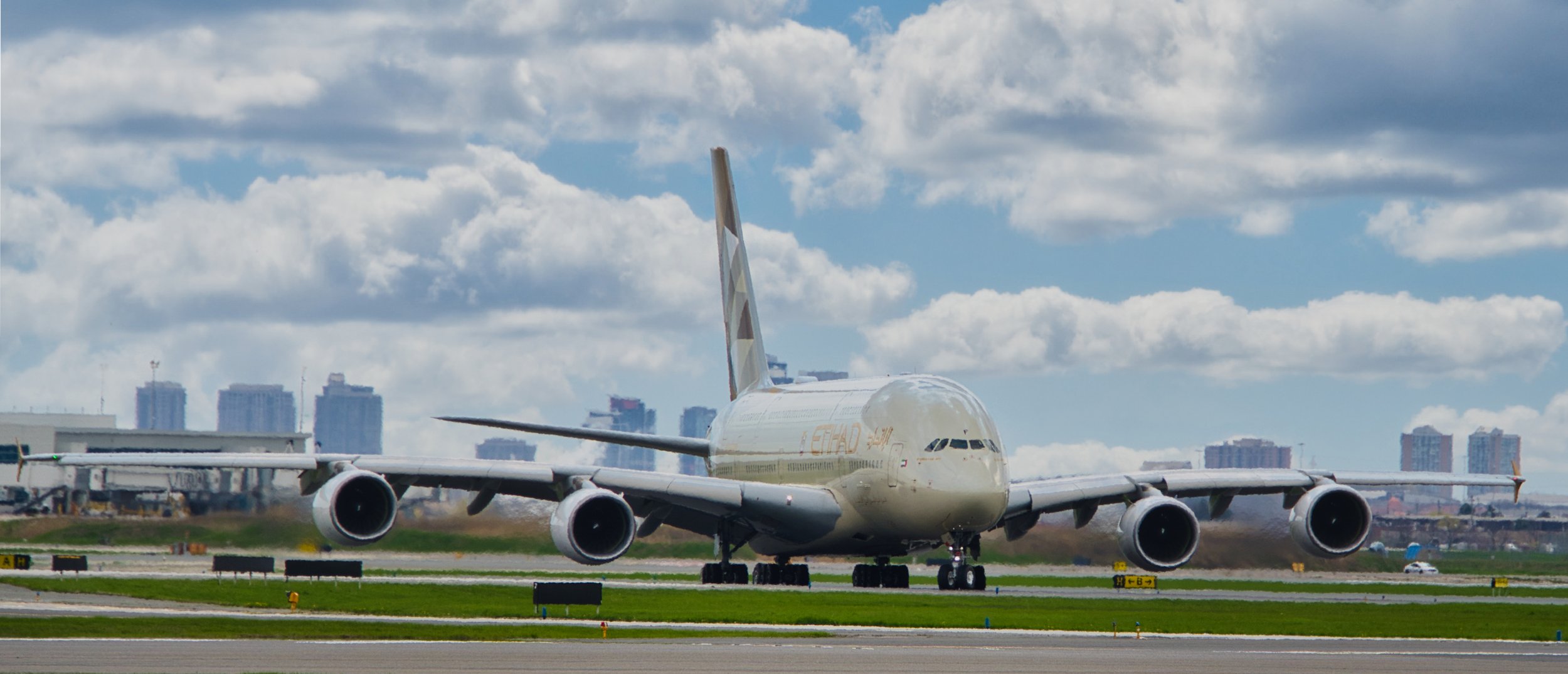 A large Etihad Airways airplane on the runway near a city skyline under a partly cloudy sky.