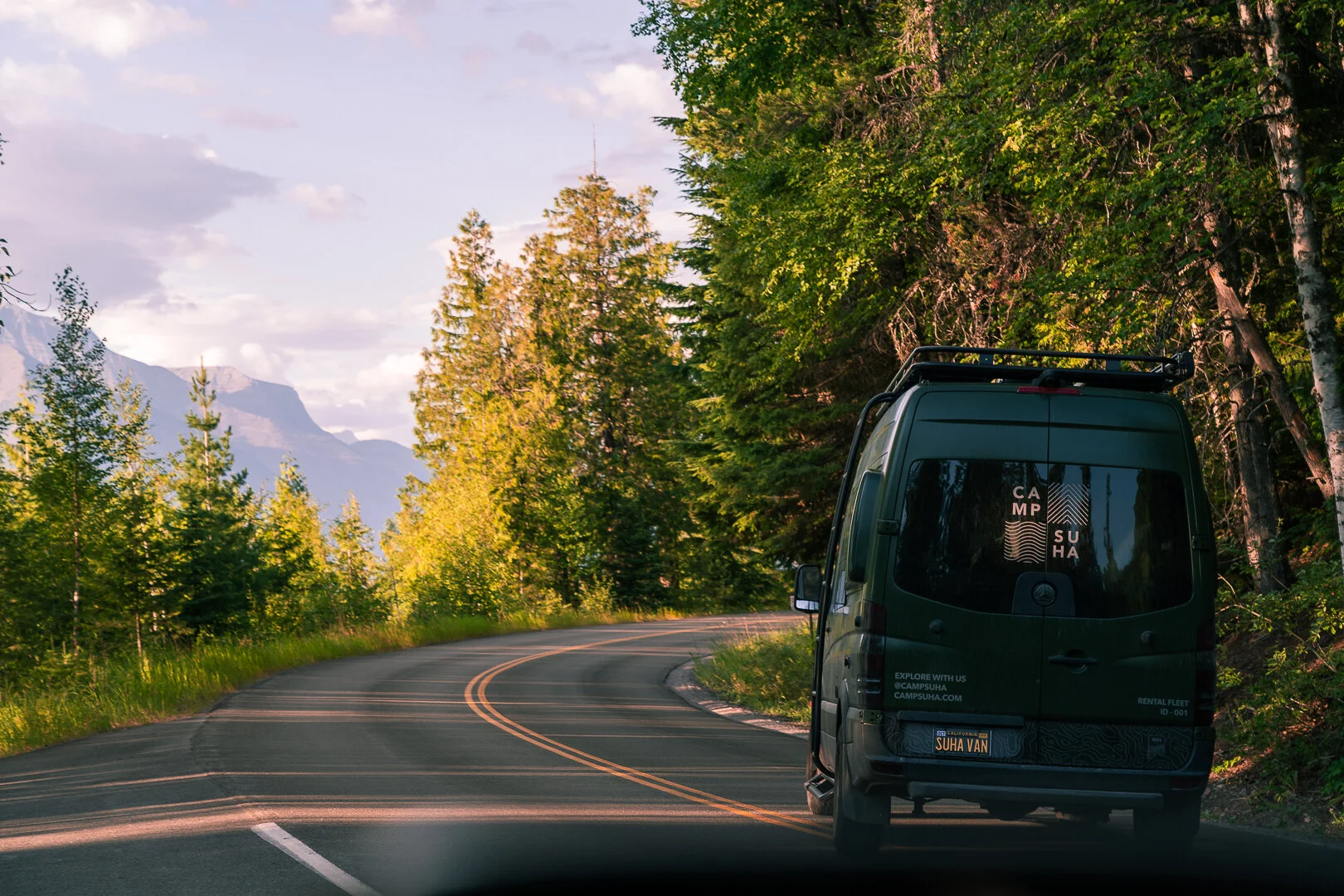 Van driving on a winding road through a forest with mountains in the background.