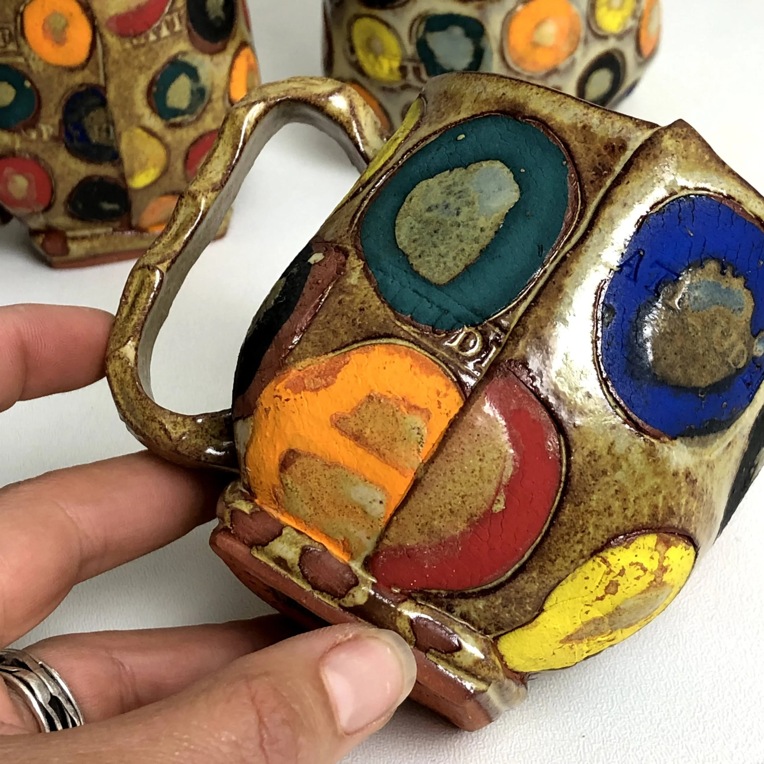 Colorful ceramic mug with abstract designs in blue, orange, red, yellow, and green, featuring a textured, glazed surface, held by a hand.