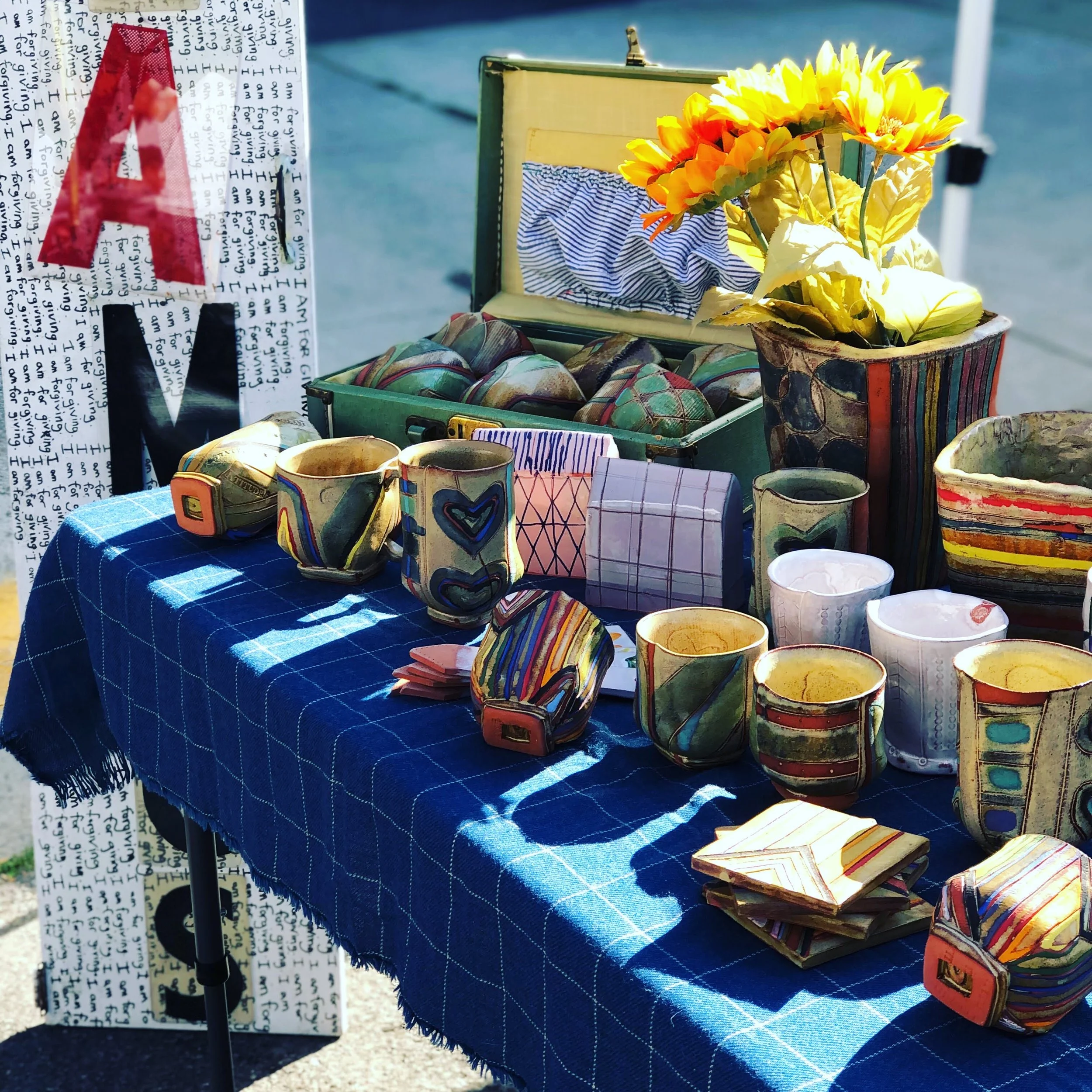 Table at an outdoor market displaying colorful pottery, cups, a jewelry box, and vibrant artificial flowers, with a decorative sign featuring the word 'LOVE' on the side.