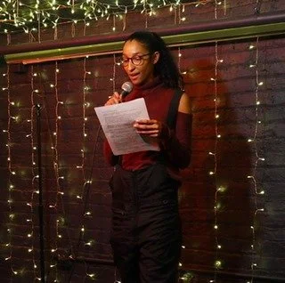 Lissette speaking into a microphone while reading from a sheet of 8x11 white paper with words printed on both sides. There are white lights hanging on the wall behind the person speaking who is the only person pictured.