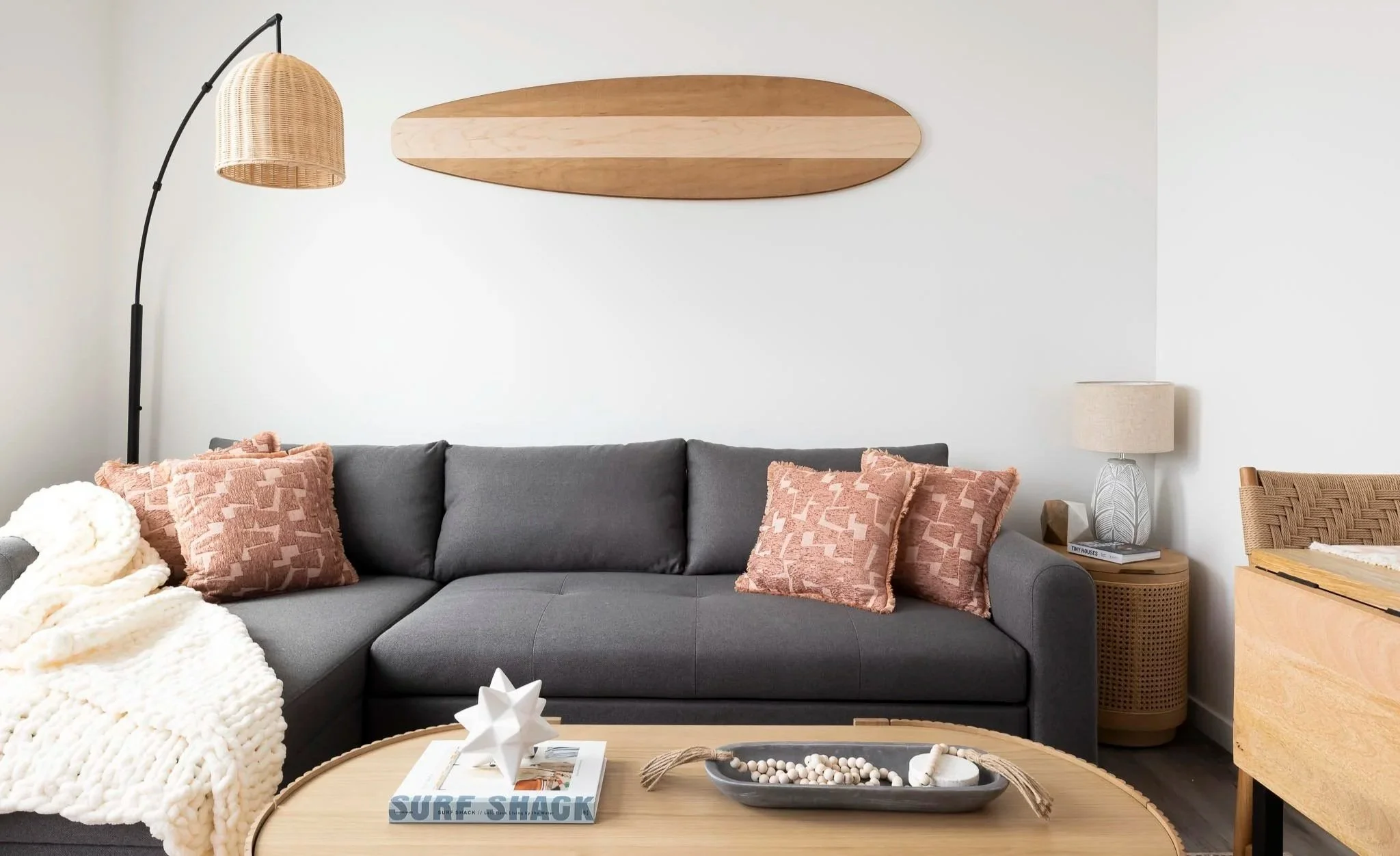 A modern living room with a gray sofa decorated with pink textured pillows, a white chunky knit blanket, a round wooden coffee table with a decorative star and a tray with beads and stones, a wicker floor lamp, a wooden side table with a textured vase lamp, and artwork resembling a surfboard hanging on a white wall.