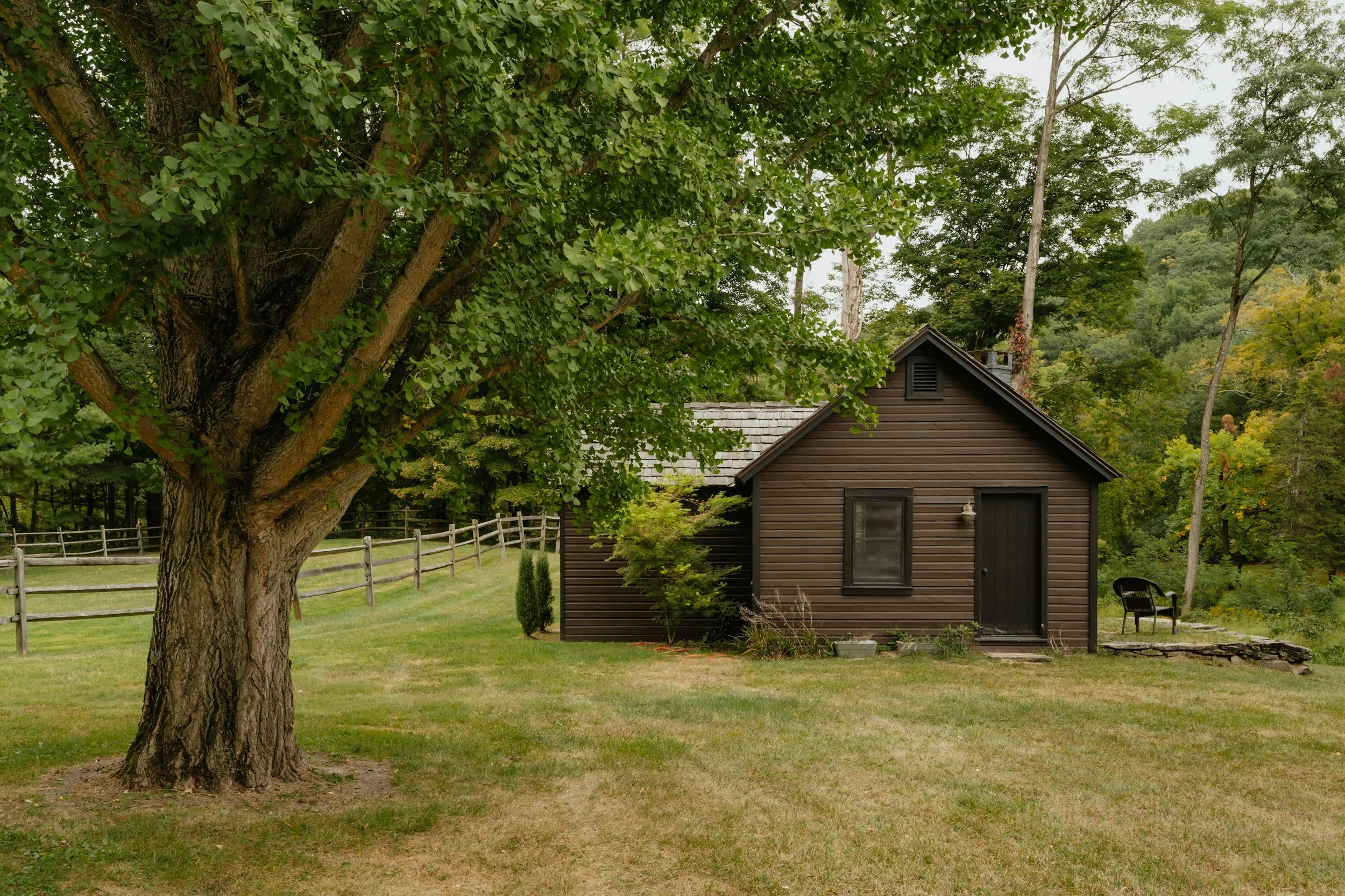 little brown cabin in the woods surround by green grass, trees and a picket fence