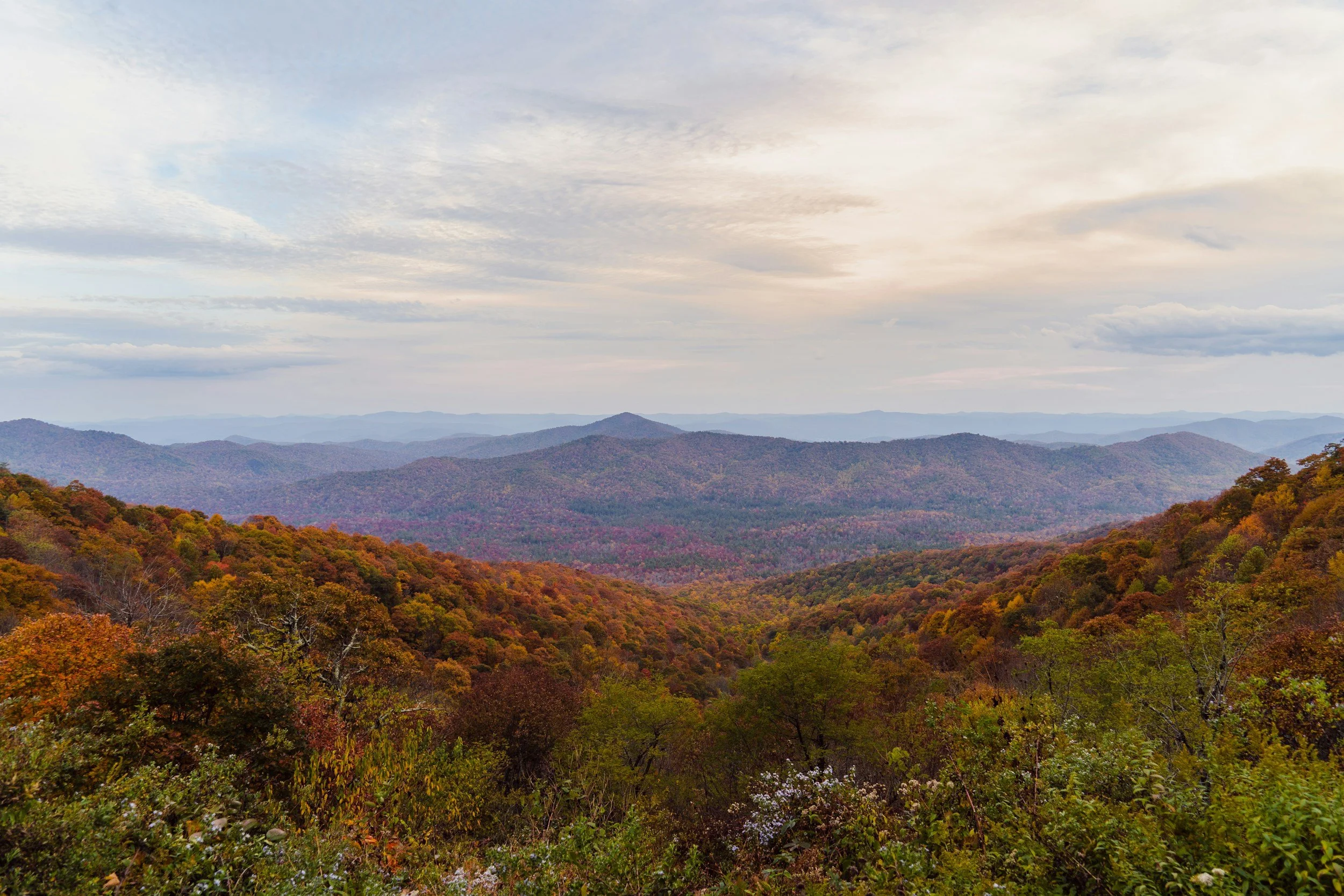 view of sun through clouds over the blue ridge mountains in the fall with shades of red orange and green.