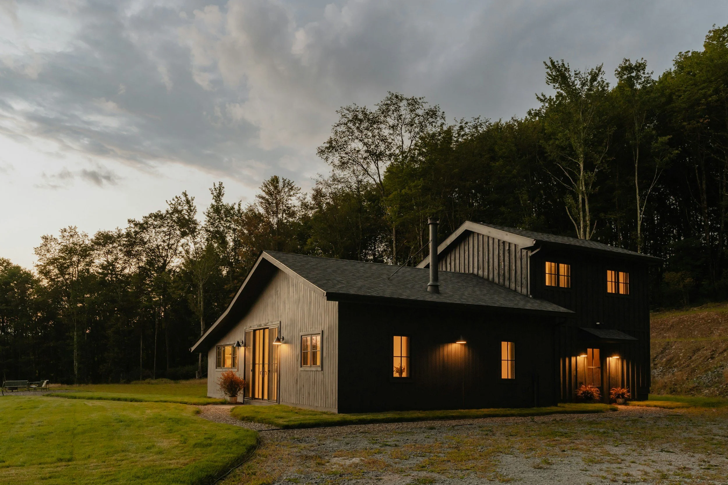 black house surrounded by woods with warmly lit windows against a darkening evening sky