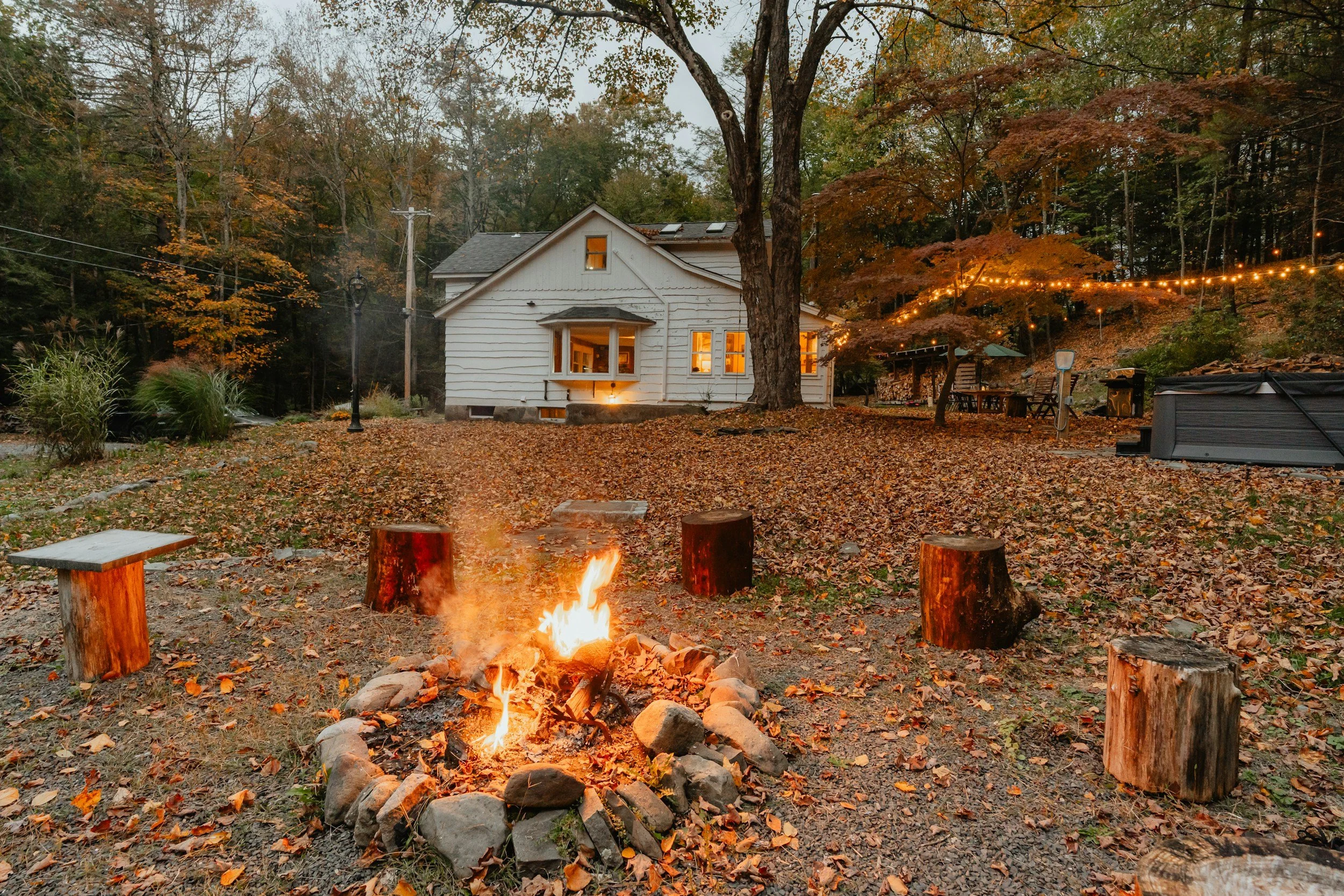 firepit with roaring fire, stump stools, and a white house in the background