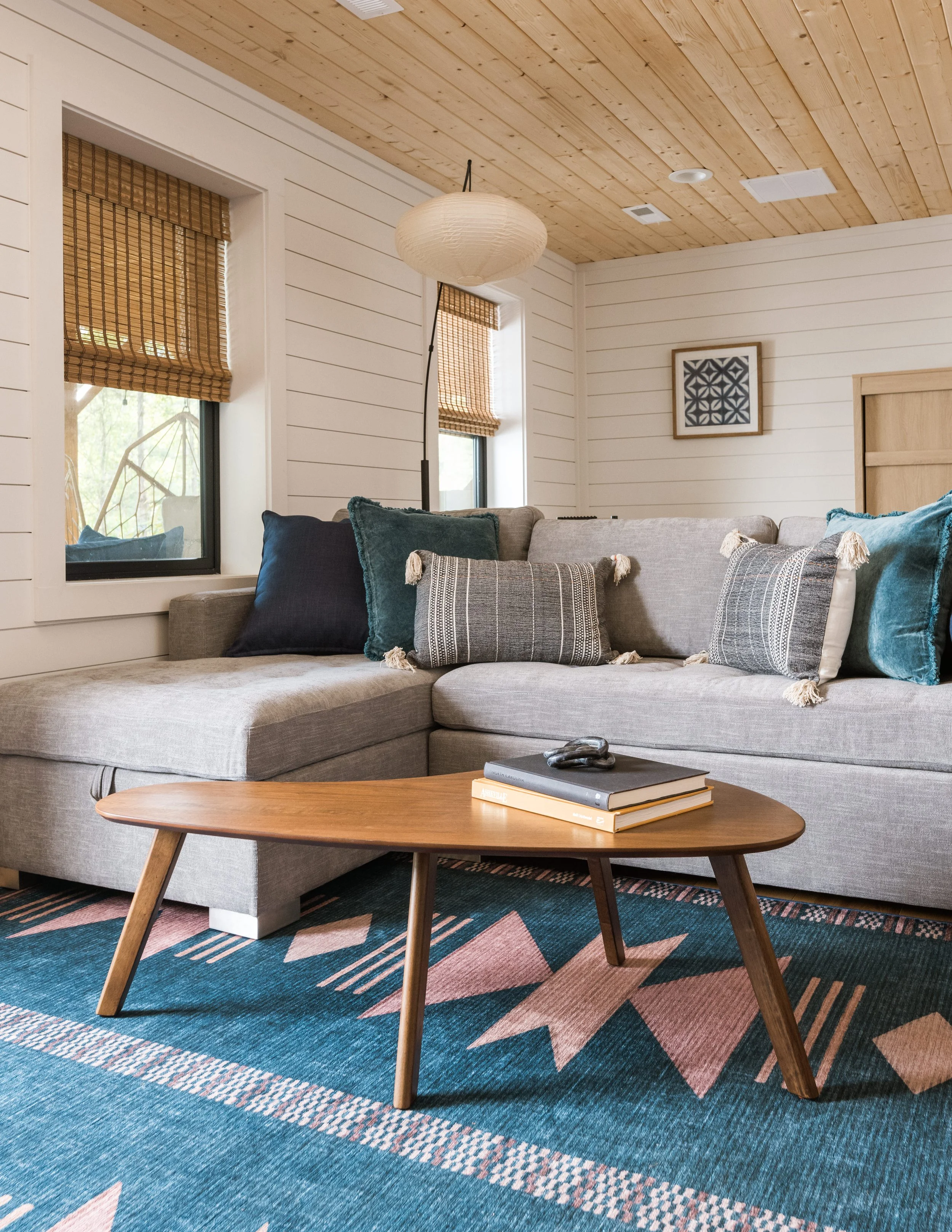 Living room with a gray sectional sofa, decorative pillows, wooden coffee table with books and a decorative object, painted wood wall, and windows with bamboo blinds.