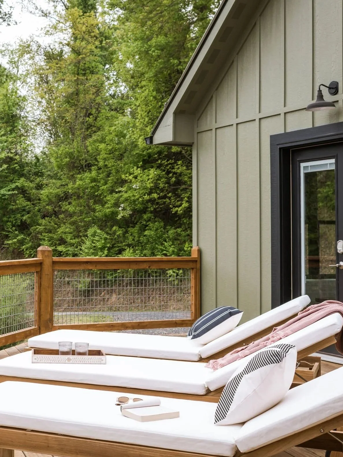 Outdoor deck with two white lounge chairs, pillows, a tray with glasses, a book, and sunglasses, surrounded by green trees.