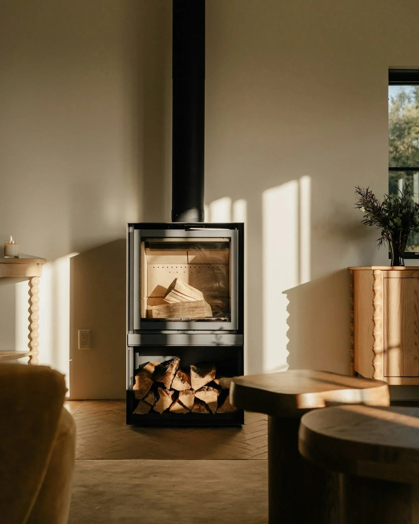 A wood stove with a glass front, stacked with firewood inside, in a living room with sunlight casting shadows on the walls, a wooden side table with a candle, and a wooden cabinet with a vase of flowers.